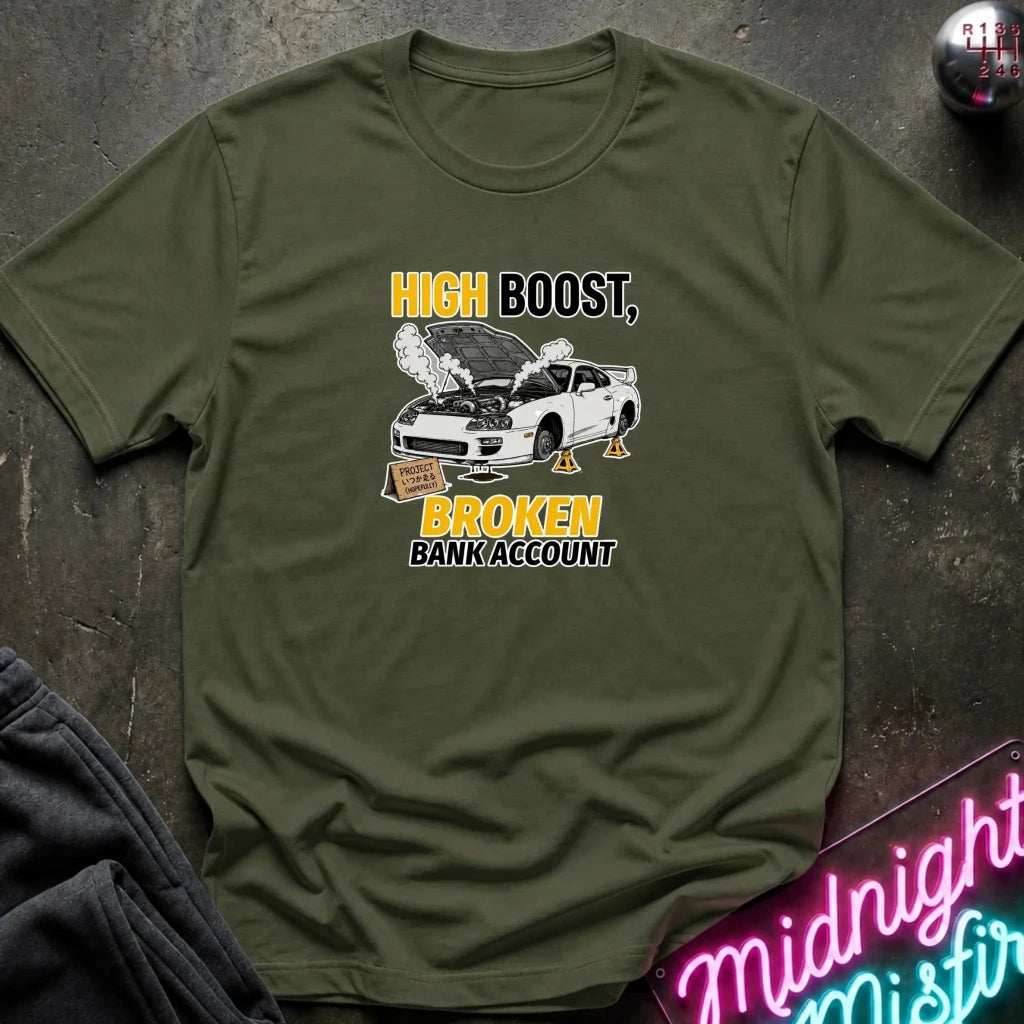 An olive green short-sleeve t-shirt with a graphic print of a white car with its hood open, surrounded by smoke, and the text 'HIGH BOOST, BROKEN BANK ACCOUNT' in yellow and black. The shirt is displayed flat on a dark textured surface with a neon sign and dark pants partially visible.