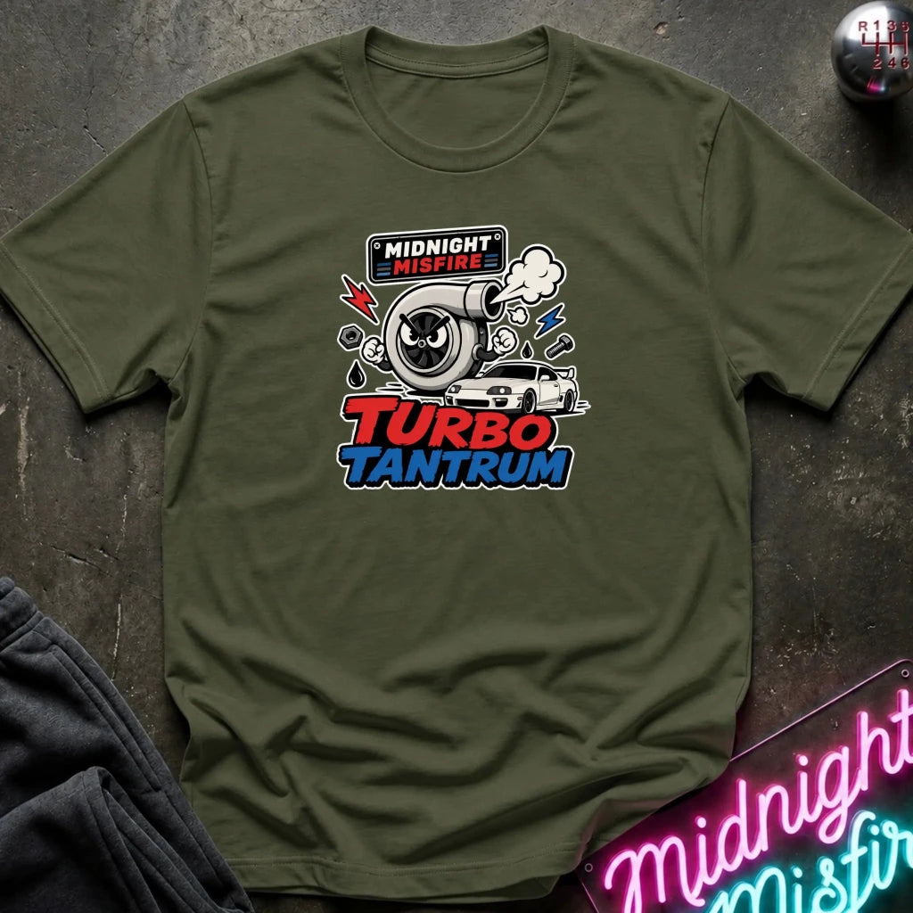 An olive green short-sleeve t-shirt with a 'Midnight Misfire Turbo Tantrum' graphic on the chest, displayed flat on a dark textured background with a neon sign and dark pants partially visible.