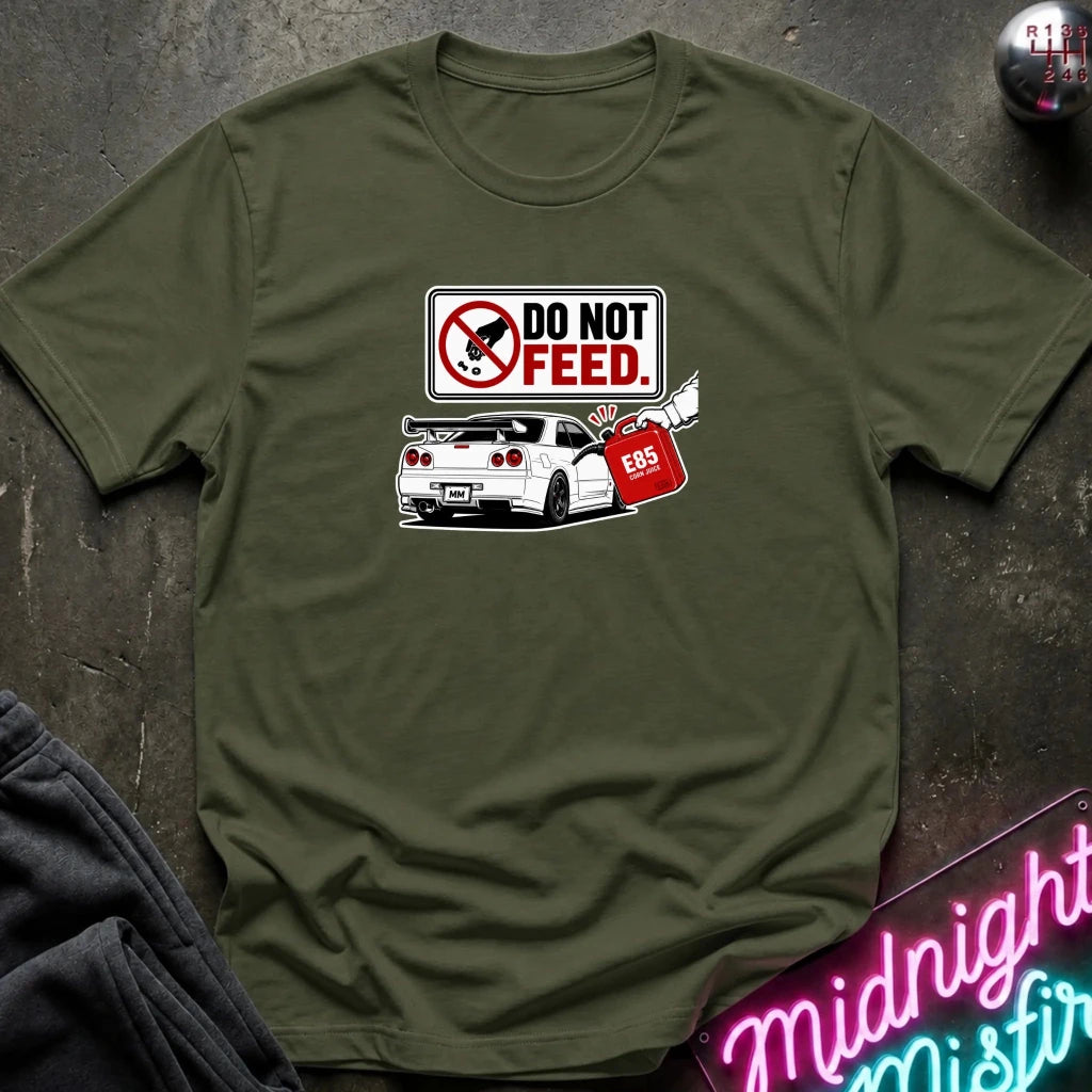 An olive green short-sleeve graphic t-shirt laid flat on a dark textured surface. The graphic features a 'DO NOT FEED' sign.