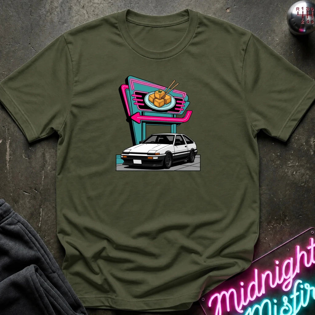 An olive green short-sleeve t-shirt laid flat on a dark textured surface, featuring a colorful graphic print of a white hatchback parked in front of a neon-style food sign. 