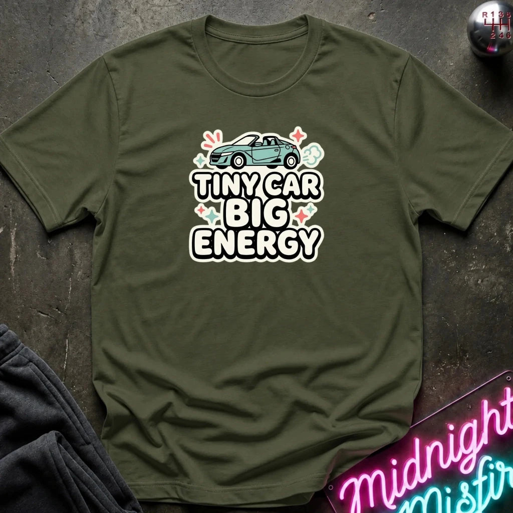 An olive green short-sleeve t-shirt laid flat on a dark textured surface, featuring a graphic print of a small blue car above the text 'TINY CAR BIG ENERGY' in white with a pink outline, surrounded by small pink and green floral accents.