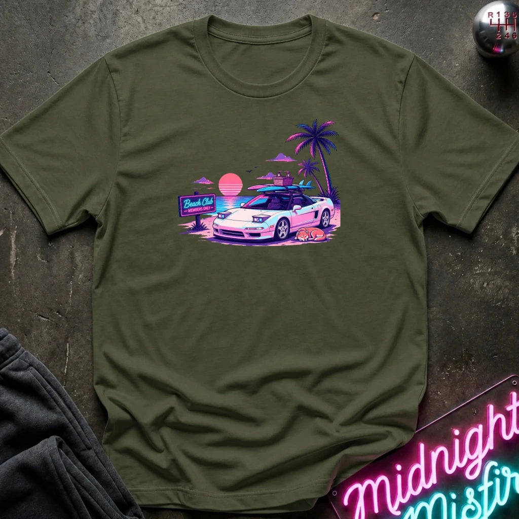 An olive green short-sleeve t-shirt laid flat on a dark textured surface, featuring a colorful retro graphic of a white sports car on a beach with palm trees and a sunset.