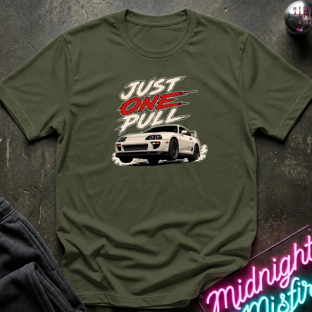An olive green short-sleeve t-shirt with a graphic print of a white sports car and the text 'JUST ONE PULL' in white and red, laid flat on a dark textured surface.
