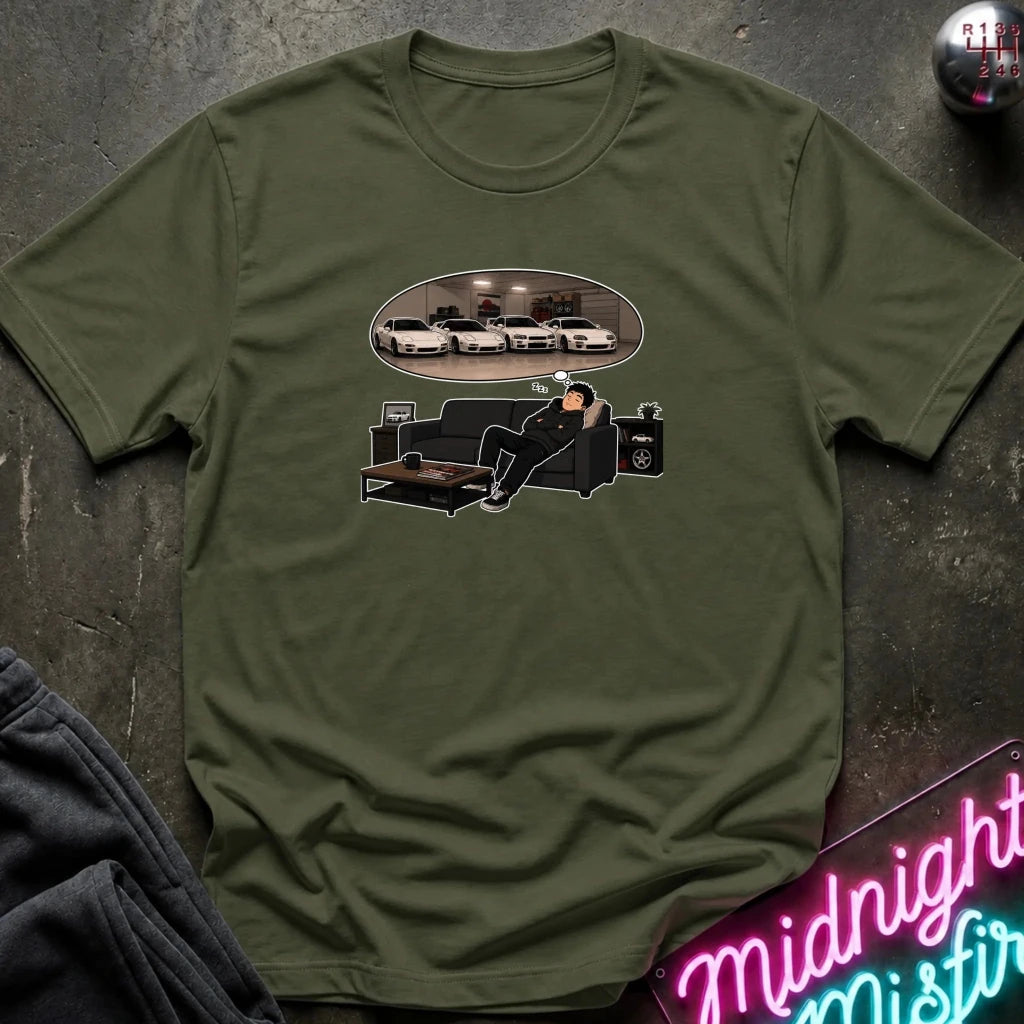 An olive green short-sleeve graphic t-shirt laid flat on a dark textured surface. The graphic shows a man sitting on a black couch dreaming, with an oval reflection of several white cars above him.