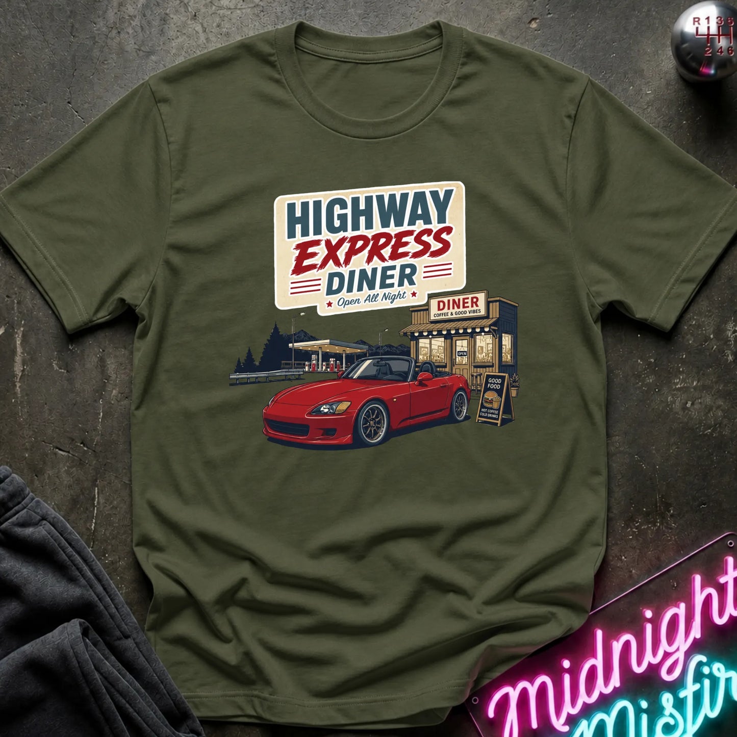 An olive green graphic t-shirt laid flat on a dark textured surface. The shirt features a retro-style print of a red convertible sports car parked outside a diner with the text 'HIGHWAY EXPRESS DINER' and 'Open 24 Hours'.