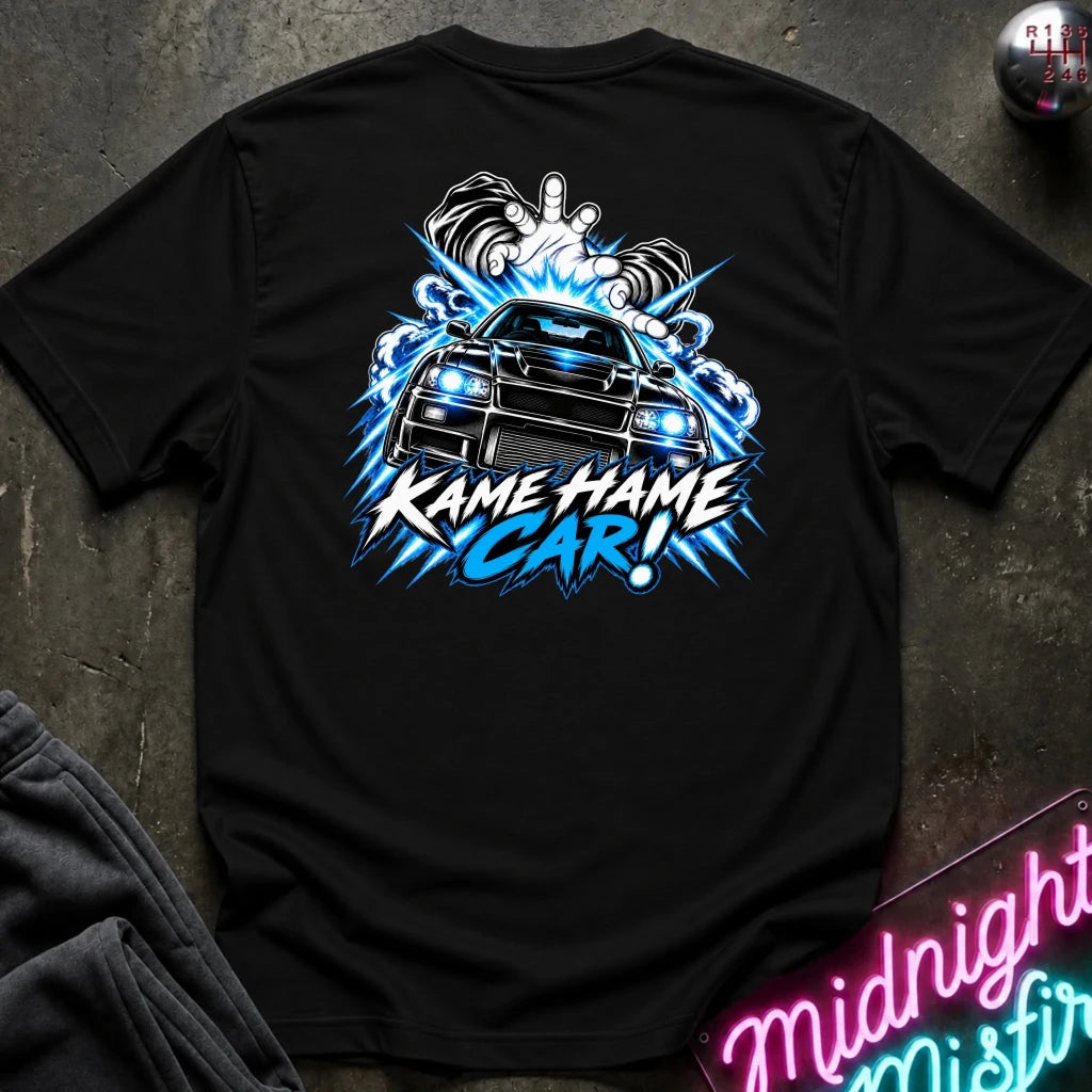 A black short-sleeve t-shirt laid flat on a dark textured surface, featuring a large back graphic of a blue and white car with the text 'KAME HAME CAR!' and a hand gesture above it. Partially visible items at the edges include dark fabric and a neon-style sign.