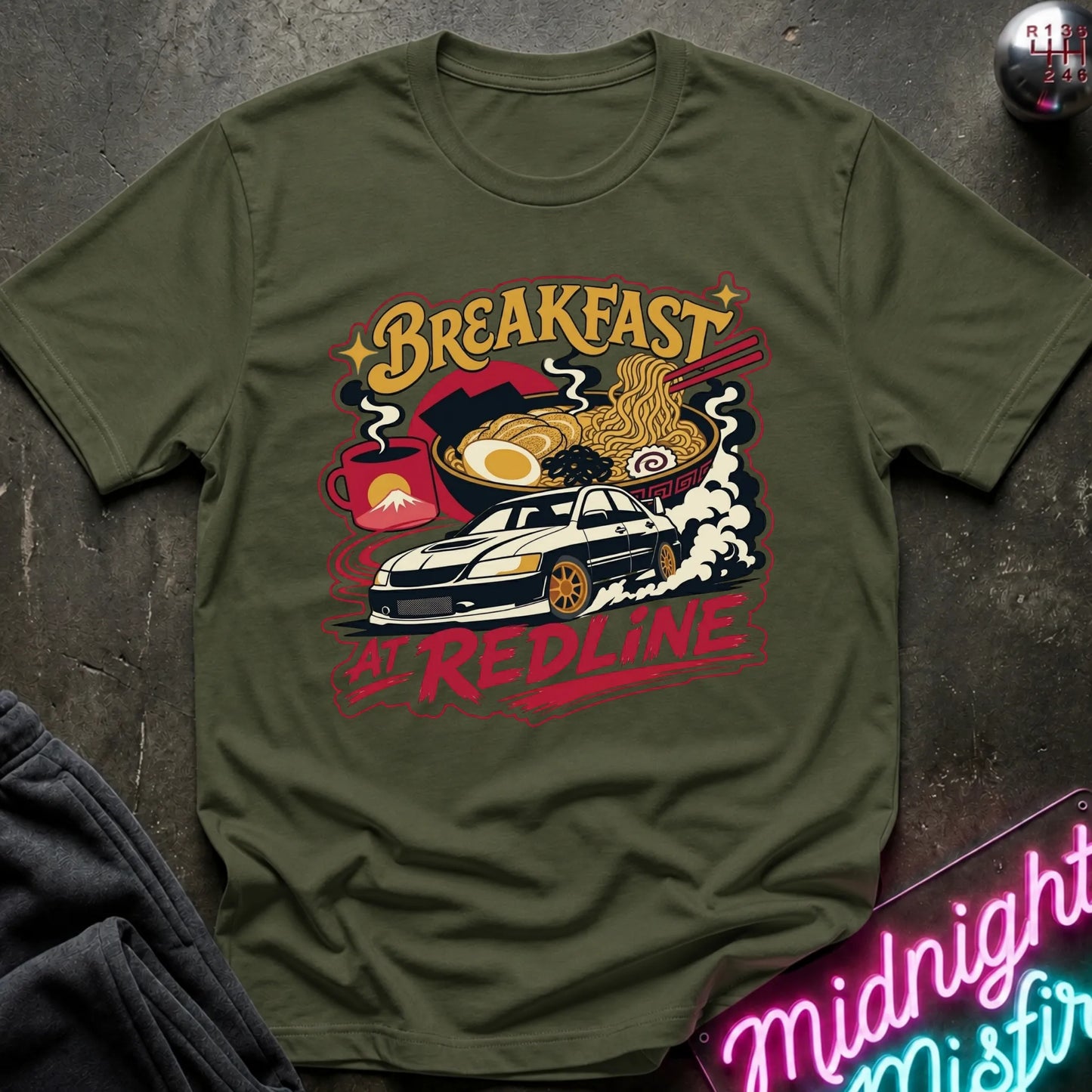 An olive green short-sleeve graphic T-shirt laid flat on a dark textured surface. The shirt features a colorful retro-style print with the words “Breakfast at Redline,” a white car, ramen noodles, an egg, and red abstract shapes.