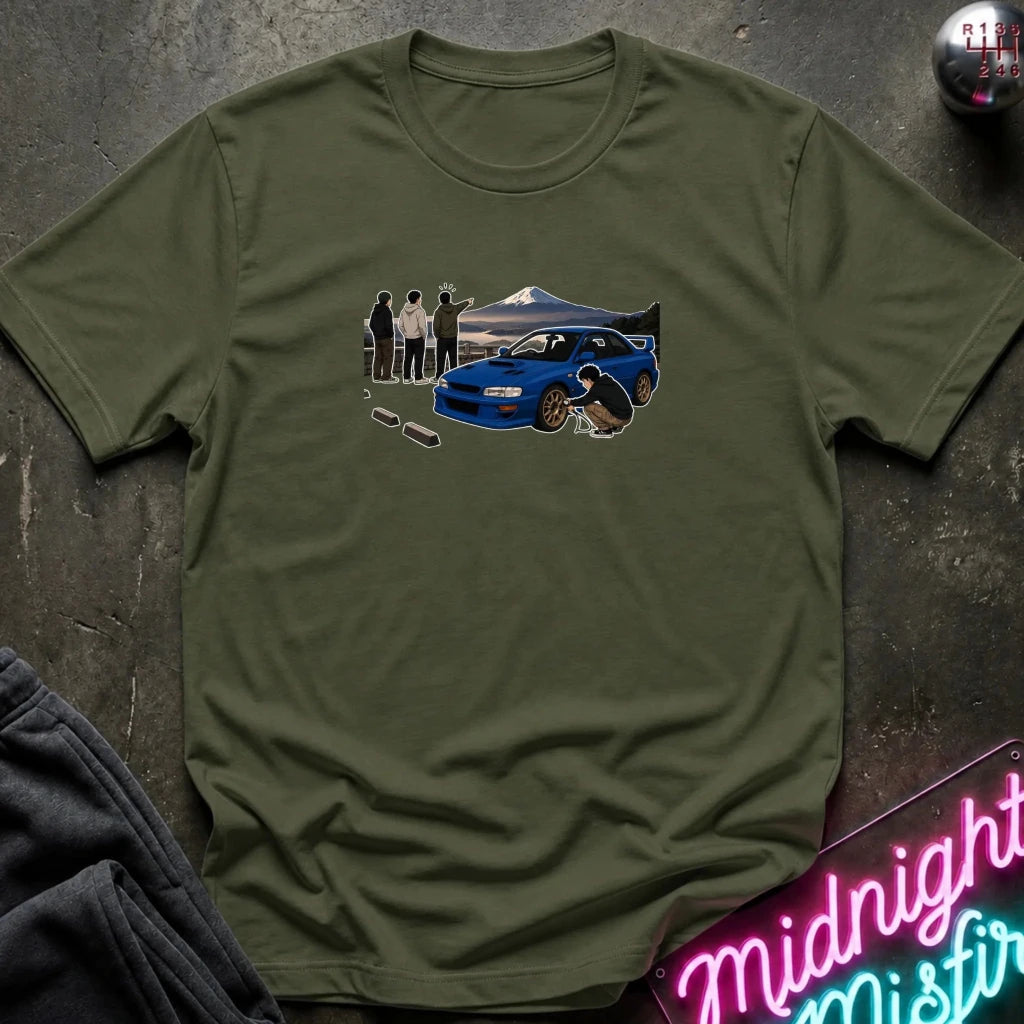 An olive green short-sleeve t-shirt laid flat on a dark textured surface, featuring a graphic print of a blue sports car, a mountain, and a group of people. A neon sign and folded dark fabric are partially visible in the corners.