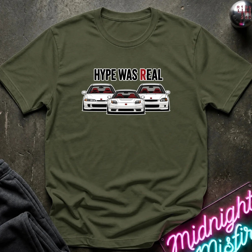 An olive green short-sleeve t-shirt laid flat on a dark textured surface. The shirt features a graphic print of three white sports cars with red interiors and the text 'HYPE WAS REAL' above them.