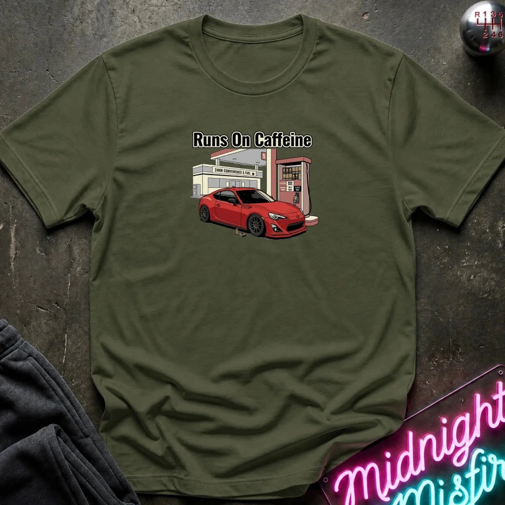 An olive green crewneck t-shirt laid flat on a dark textured surface, featuring a graphic of a red sports car at a gas station with the text 'Runs On Caffeine'.