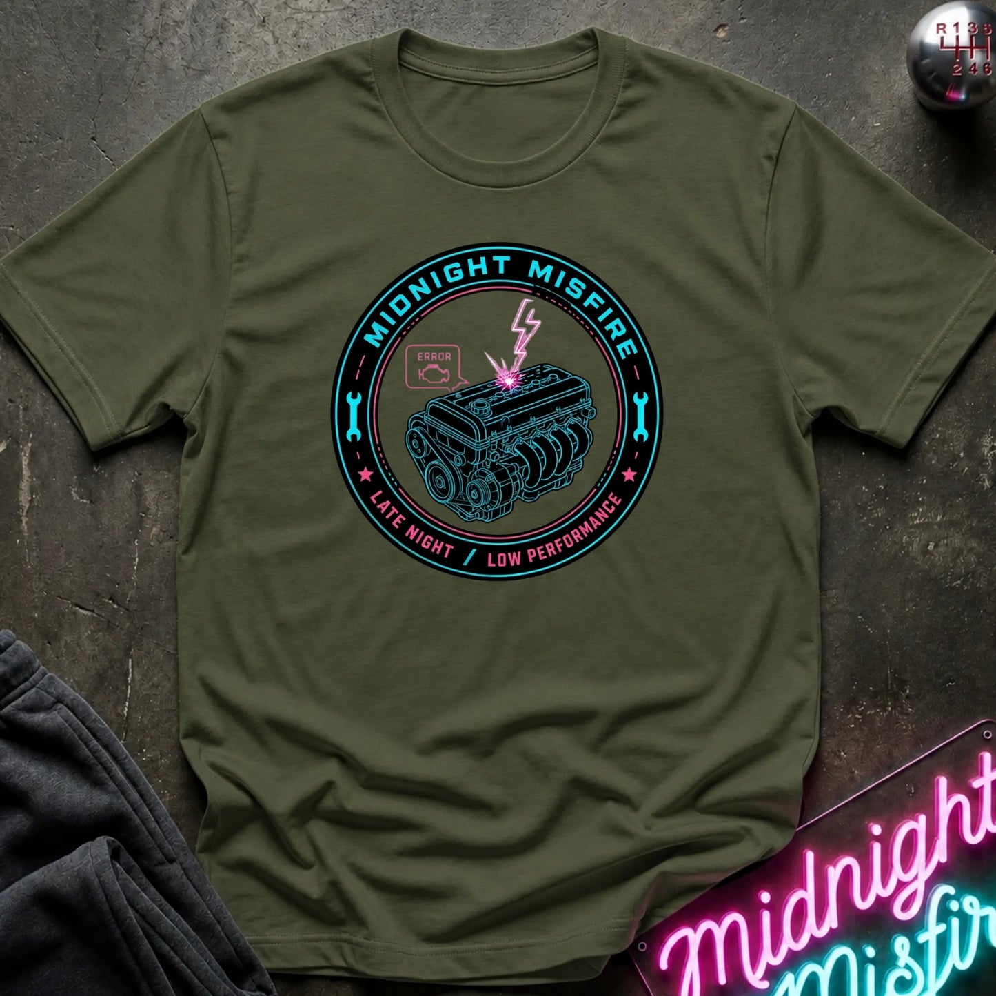 An olive green crewneck t-shirt with a circular graphic on the chest reading 'Midnight Misfire' and 'Late Night / Low Performance' around a tank illustration with a lightning bolt. The shirt is laid flat on a dark textured surface with a neon sign and dark pants partially visible.