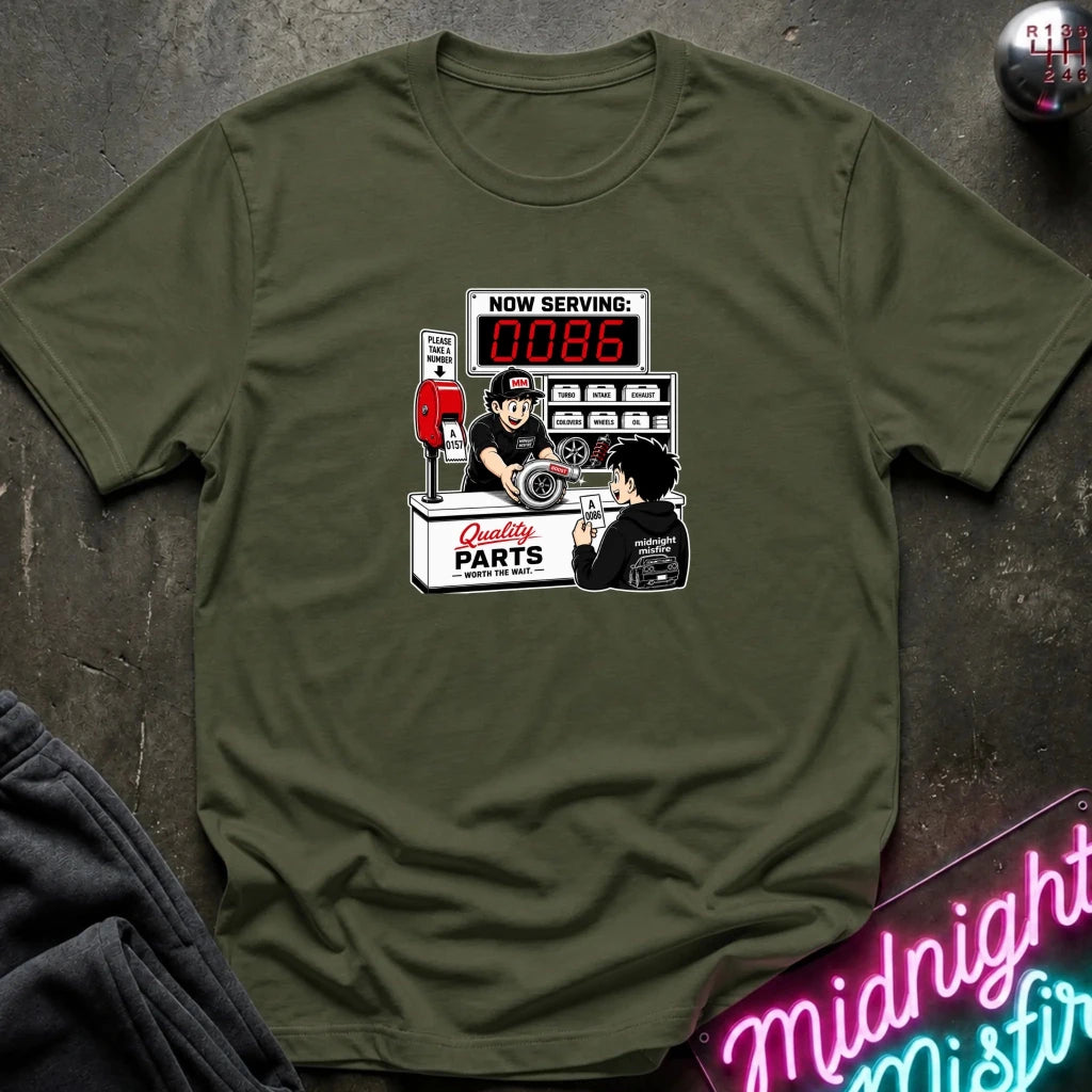 An olive green crewneck t-shirt with a retro-style graphic of a garage mechanic counter, displayed on a dark textured background with a neon sign and clothing prop.