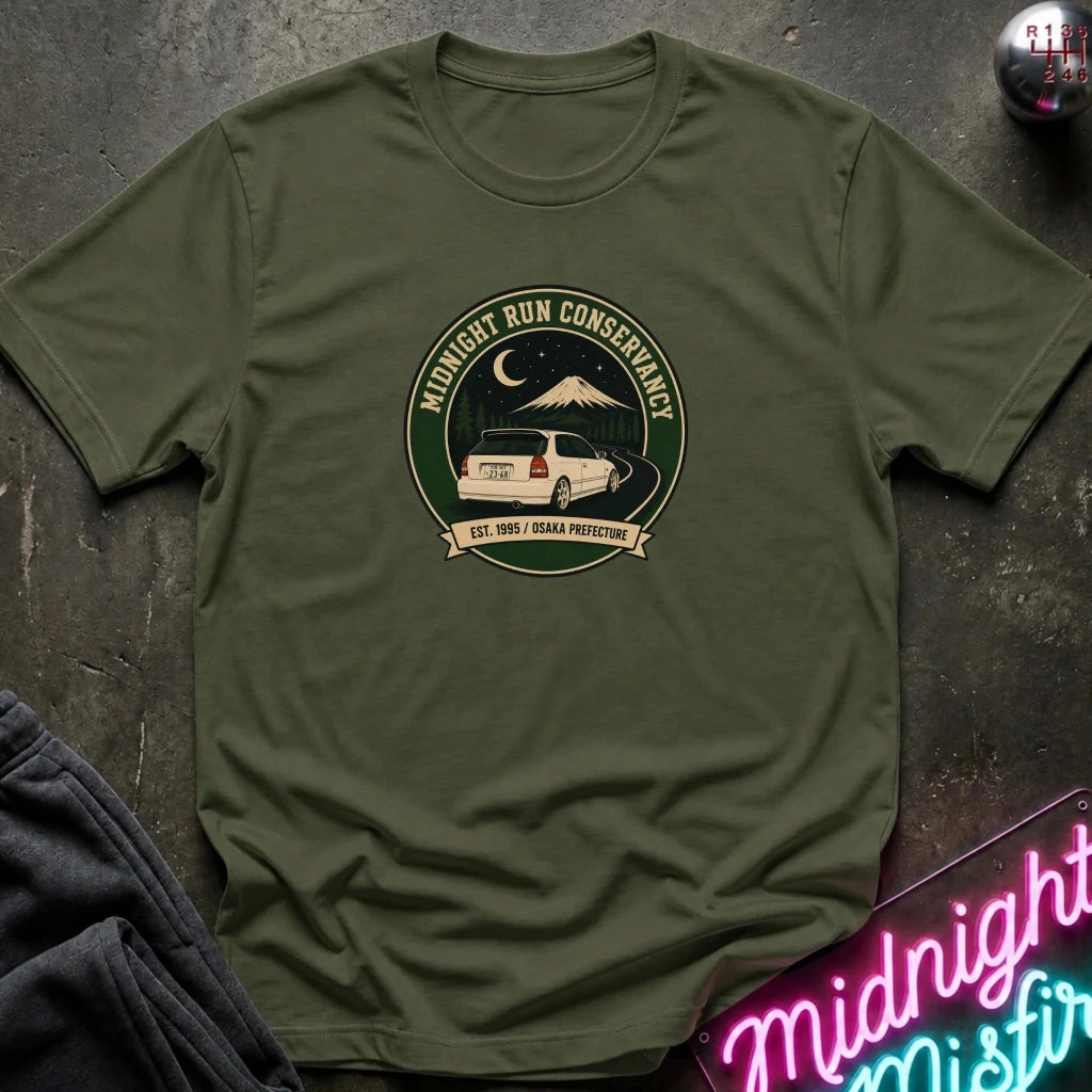 An olive green short-sleeve t-shirt with a circular graphic on the chest reading 'Midnight Run Conservancy' and showing a white vehicle, mountain, and moon. The shirt is laid flat on a dark textured surface with a neon sign and dark pants partially visible.