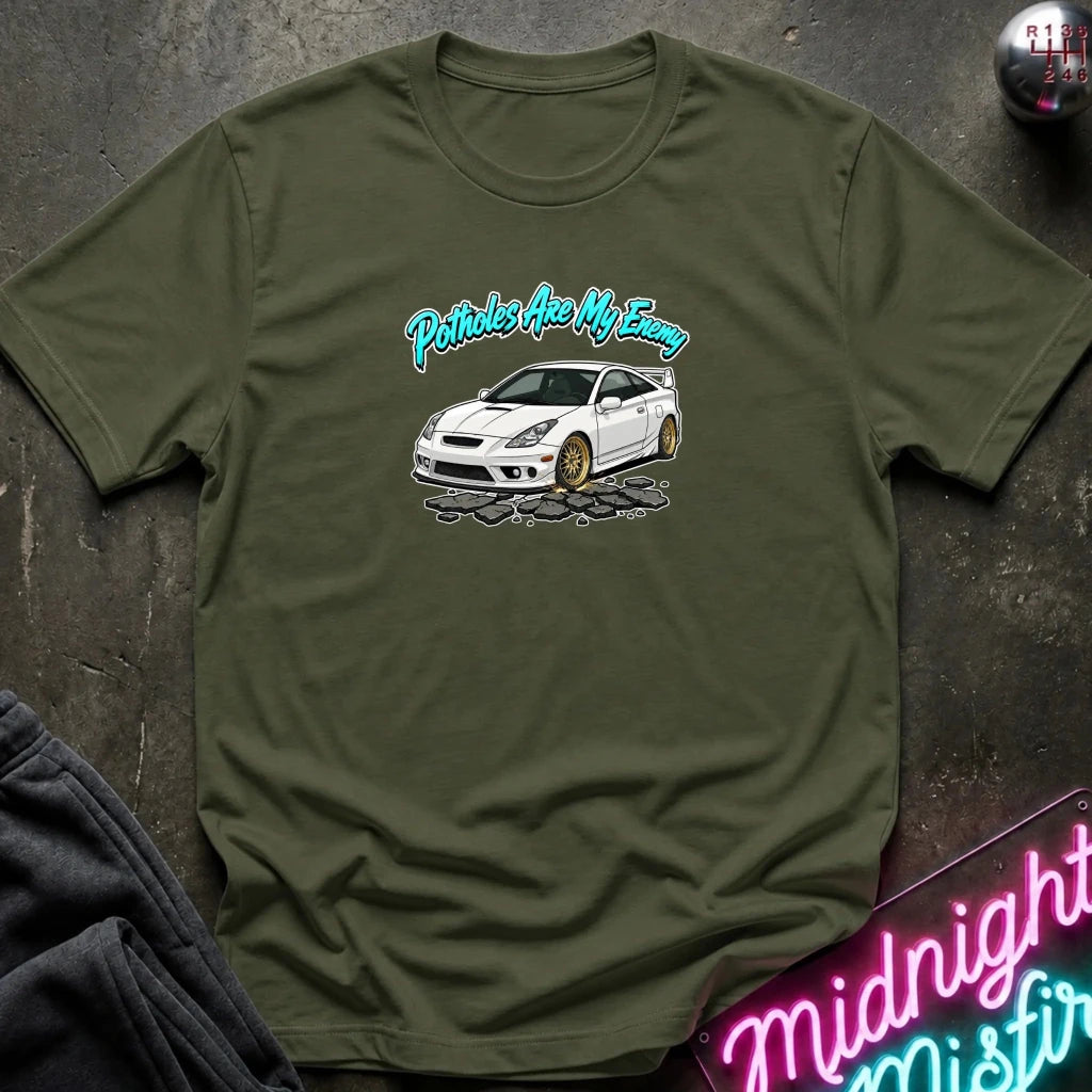 An olive green short-sleeve t-shirt laid flat on a dark textured surface. The shirt features a graphic print of a white sports car with gold wheels and the text 'Potholes Are My Enemy' in a teal script.