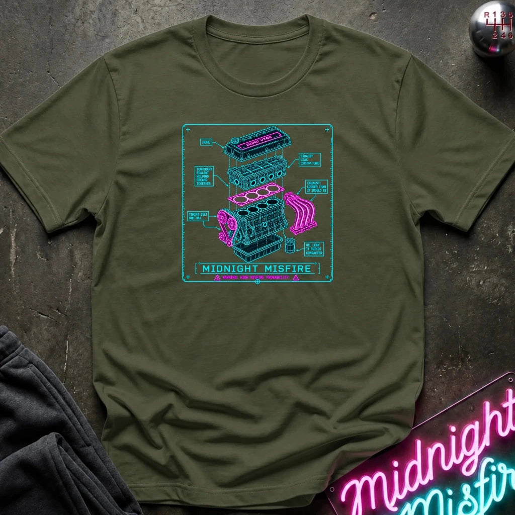 olive short-sleeve graphic T-shirt laid flat on a dark textured surface. The shirt features a centered rectangular print with a light blue and pink exploded-style engine diagram and the text 'MIDNIGHT MISFIRE.