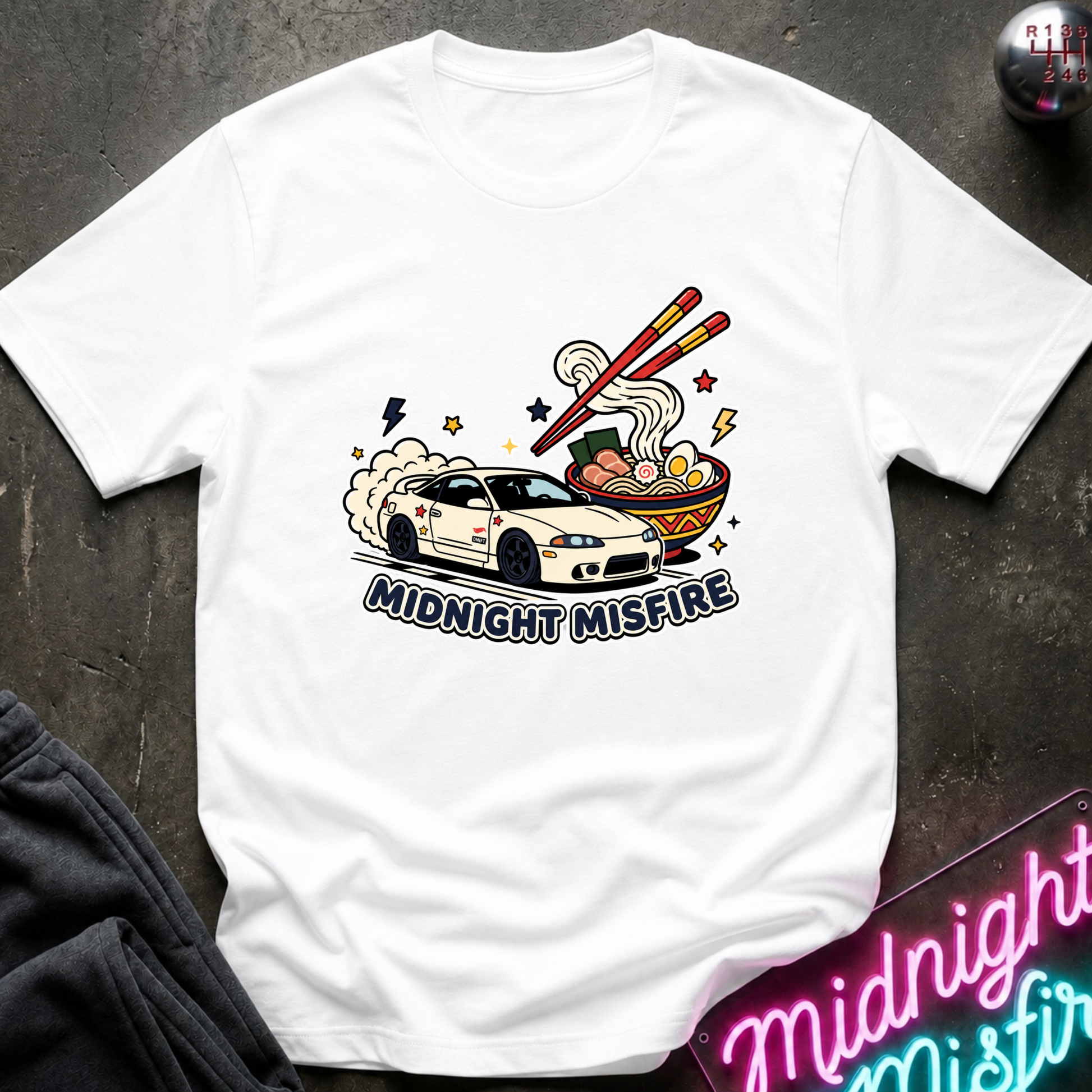 A white short-sleeve graphic t-shirt laid flat on a dark textured surface. The graphic features a white sports car with smoke, chopsticks, ramen noodles, and the text 'MIDNIGHT MISFIRE'.