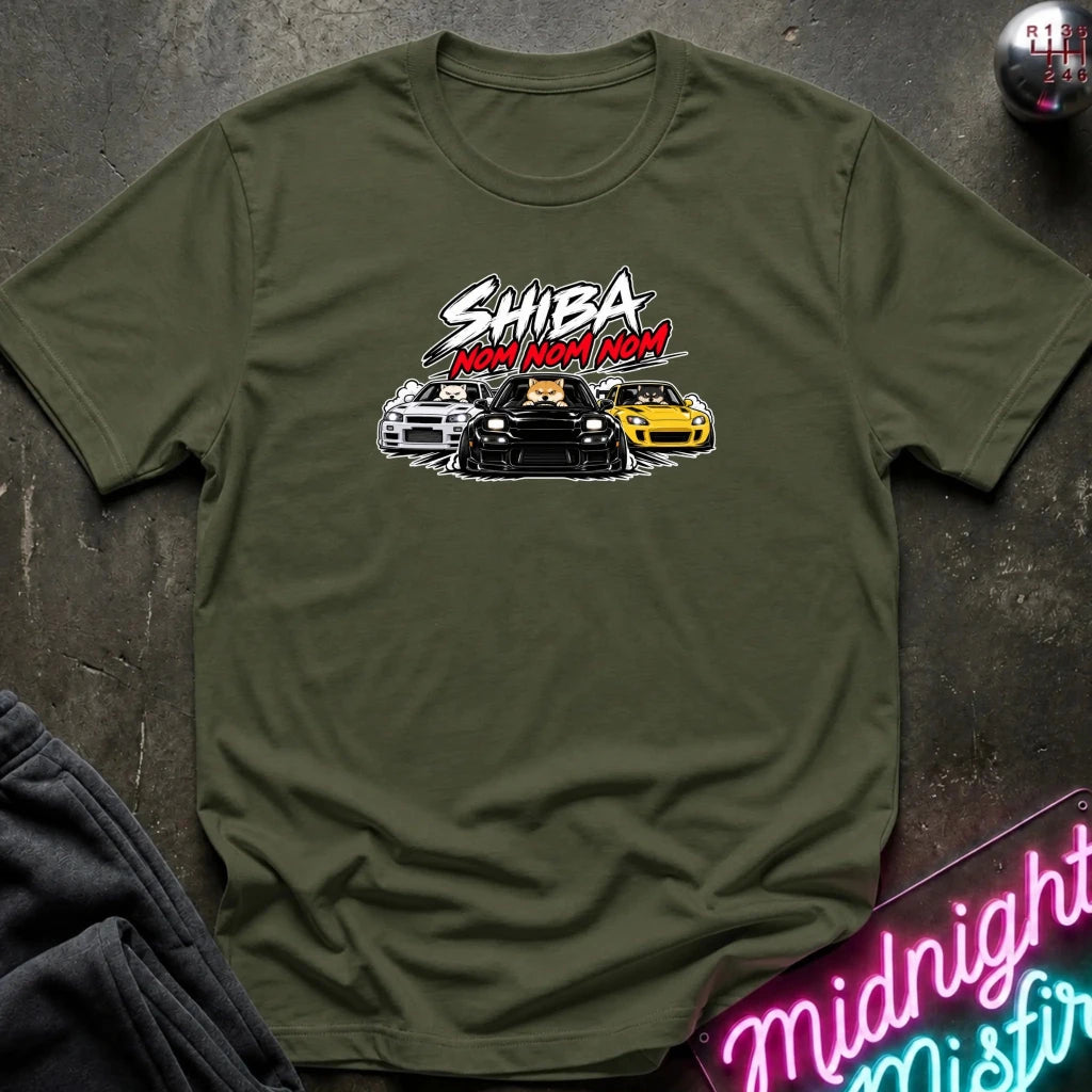 An olive green short-sleeve graphic t-shirt laid flat on a dark textured surface. The graphic features the text 'SHIBA NOM NOM NOM' in white and red, above an illustration of three cars (white, black, and yellow).
