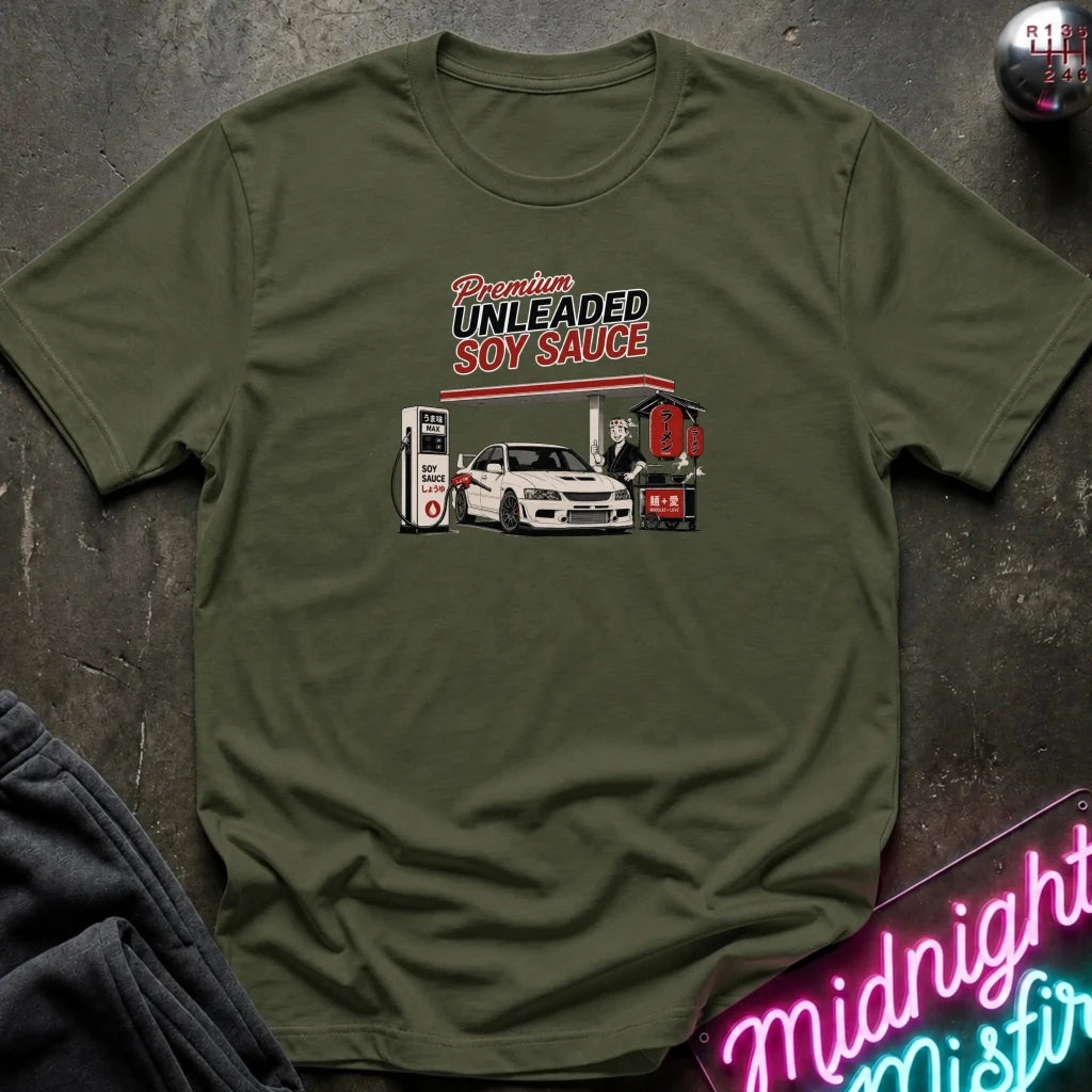 An olive green short-sleeve t-shirt laid flat on a dark textured surface, featuring a graphic print of a white sports car at a gas station with the text 'Premium UNLEADED SOY SAUCE'.
