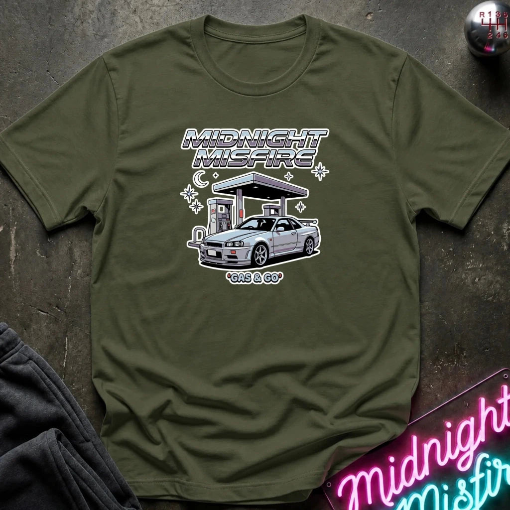 An olive green short-sleeve t-shirt with a graphic print of a silver Nissan S13 'Kits & Co' car at a gas station under the text 'MIDNIGHT MISFIRE' and 'gas & GO', laid flat on a dark textured surface.