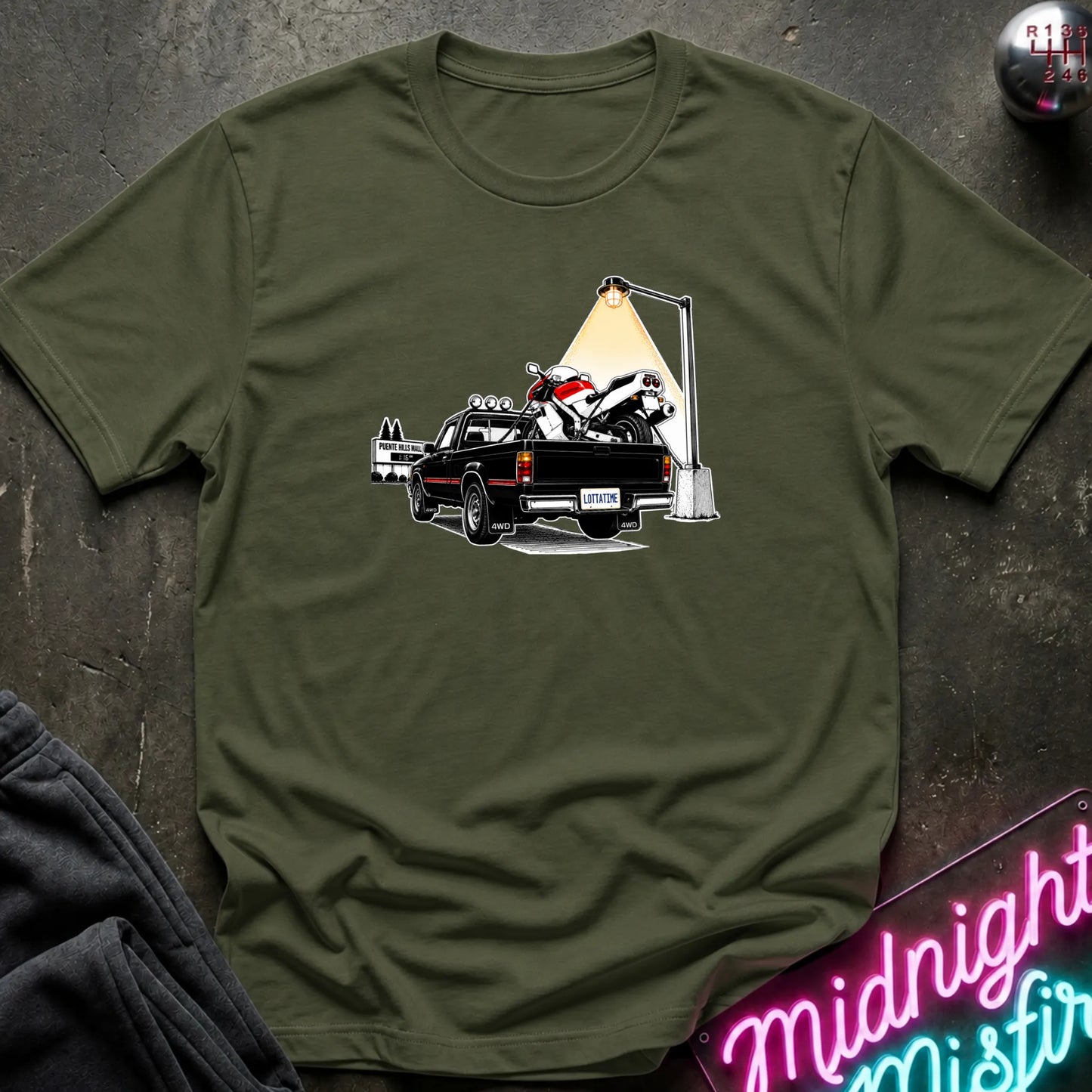 An olive green short-sleeve crewneck t-shirt featuring a graphic print of a black pickup truck carrying a white and red motorcycle under a street lamp, laid flat on a dark textured surface.