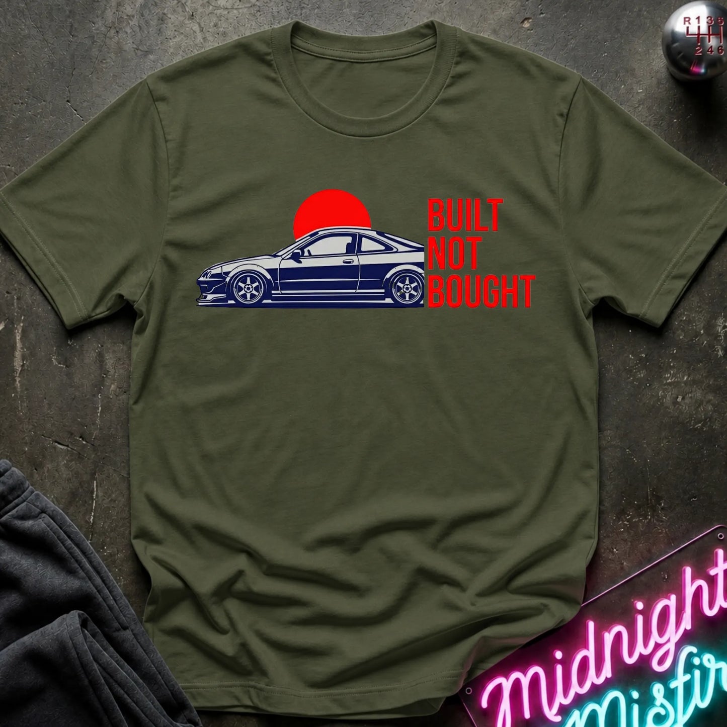 An olive green short-sleeve t-shirt laid flat on a dark textured surface. The shirt features a graphic print of a blue sports coupe car in front of a red sun, with the text 'BUILT NOT BOUGHT' printed in red to the right.
