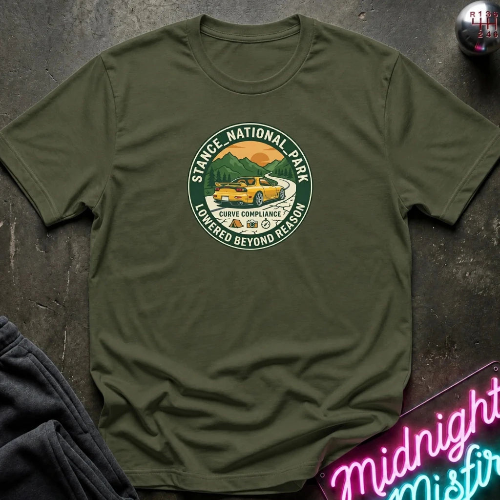 An olive green short-sleeve t-shirt laid flat on a dark textured surface, featuring a circular graphic print of a yellow car in a scenic mountain landscape with the text 'STACE NATIONAL PARK' and 'LOWEDGED BEYOND REASON'.