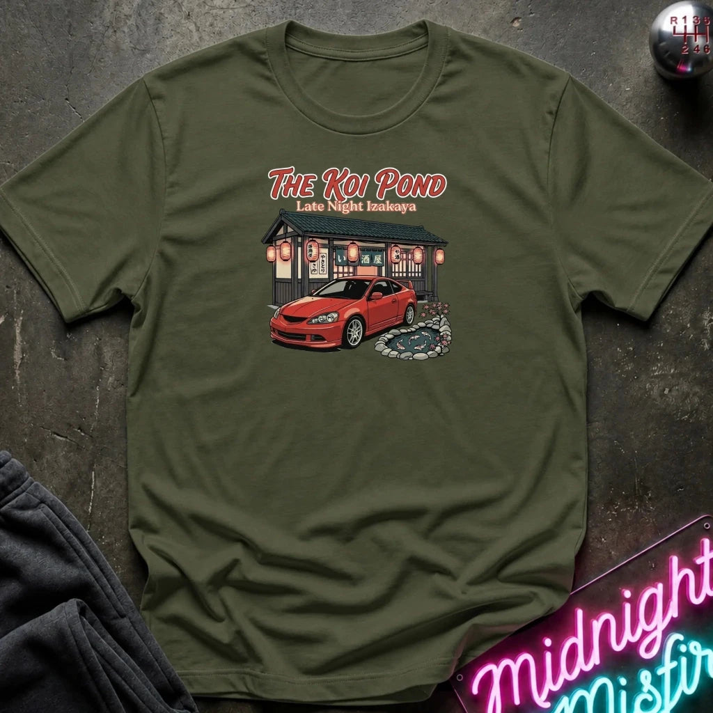 An olive green short-sleeve graphic t-shirt laid flat on a dark textured surface. The graphic features the text 'THE KOI POND Late Night Hamburger' above an illustration of a red sports car parked in front of a restaurant with red lanterns and a koi pond.