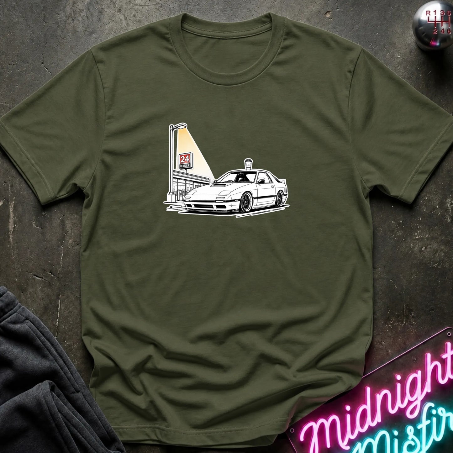 An olive green short-sleeve crewneck t-shirt laid flat on a dark textured surface, featuring a black line art graphic of a white sports car parked next to a lit street lamp.