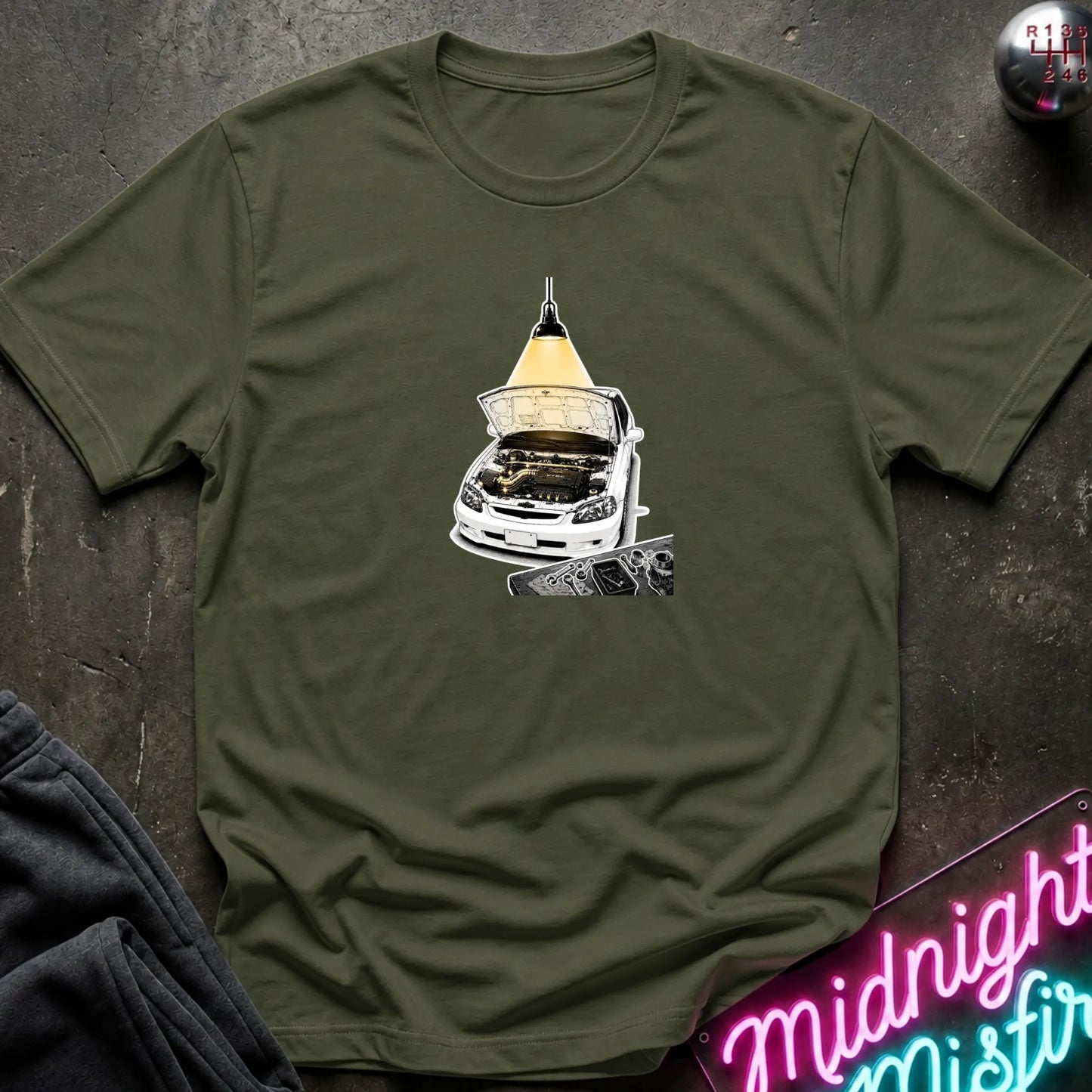 An olive green short-sleeve t-shirt laid flat on a dark textured surface, featuring a graphic print of a white car with its hood open under a glowing yellow lamp. 