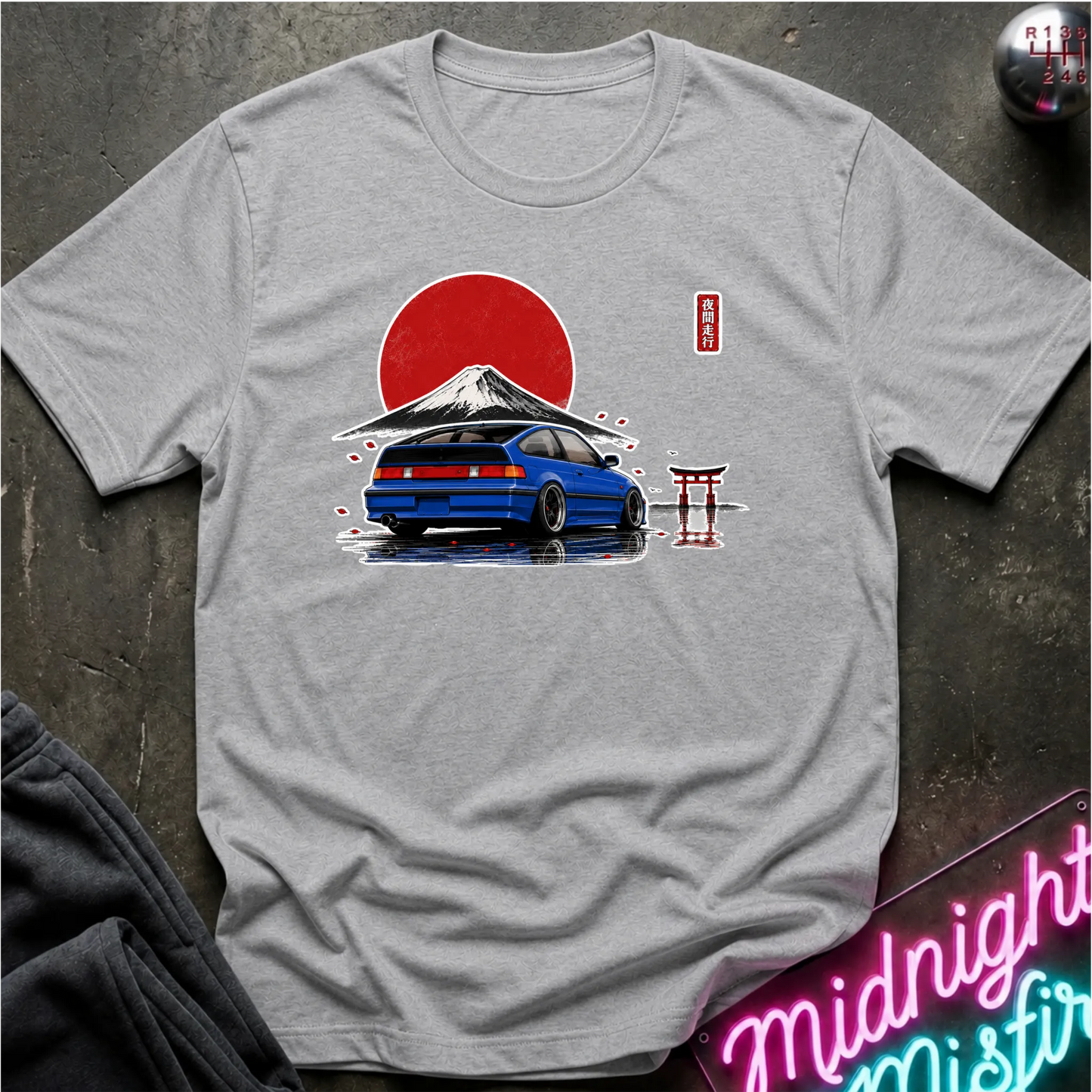A light gray crewneck t-shirt featuring a graphic print of a blue sports car in front of a red sun and Mount Fuji, styled flat on a dark textured background with a neon sign and dark pants partially visible.