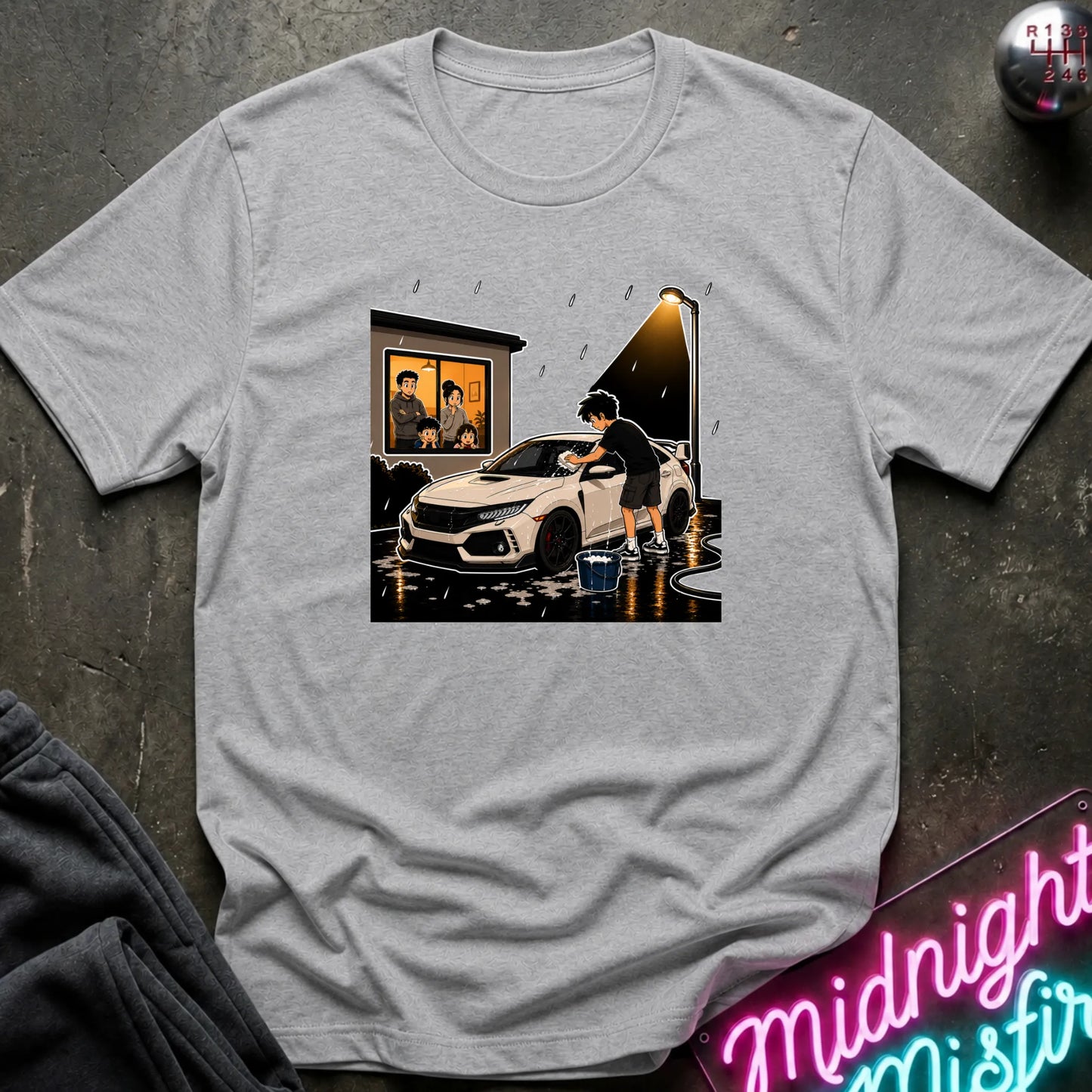 A light gray heather crewneck t-shirt featuring a graphic print of a person washing a white sports car in the rain, styled flat on a dark textured background with a neon sign and jacket in the corners.