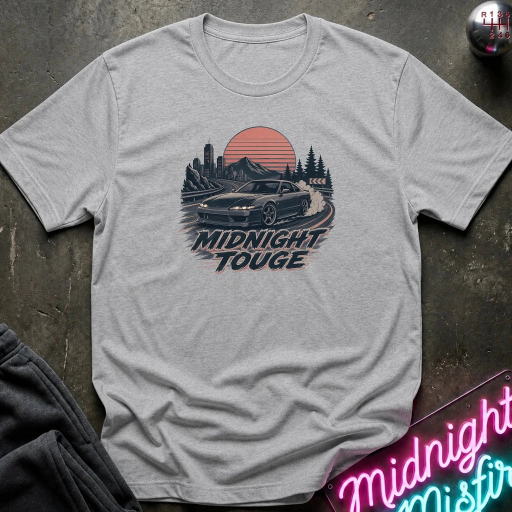 A light gray heather crewneck t-shirt laid flat on a dark textured surface. The shirt features a retro-style graphic of a black sports car drifting on a road at sunset, with the text 'MIDNIGHT TOUGHE' printed below.