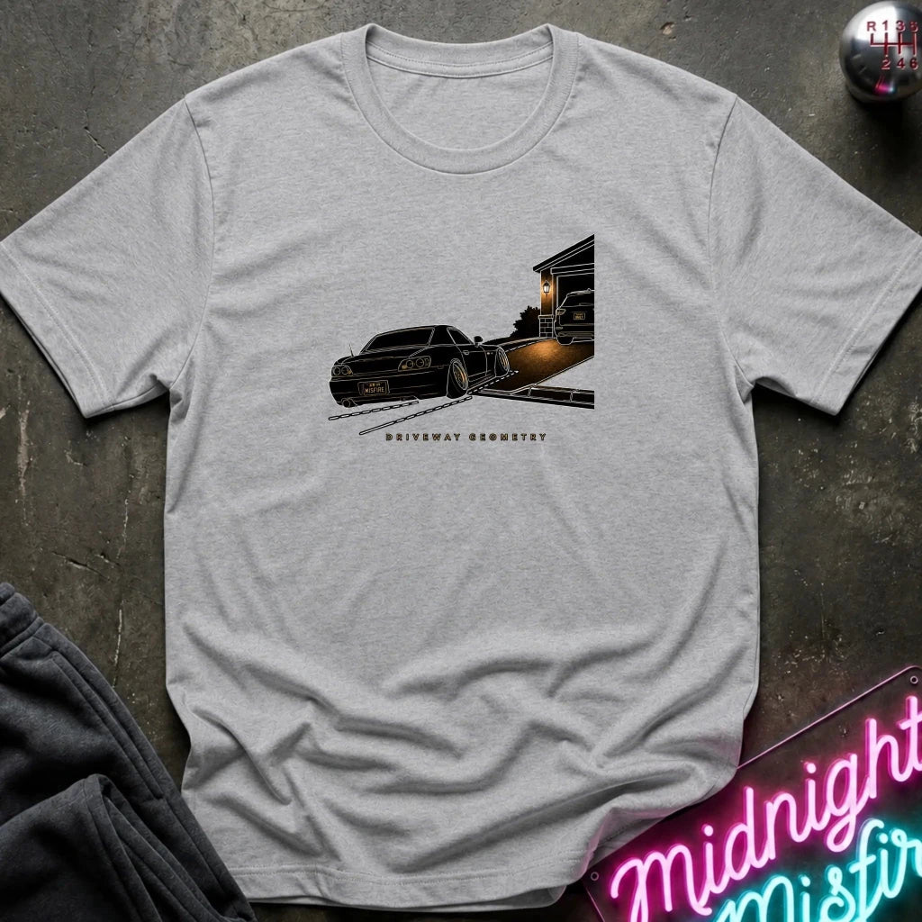 A light heather gray short-sleeve t-shirt laid flat on a dark textured surface, featuring a graphic print of a black classic car going up the driveway.