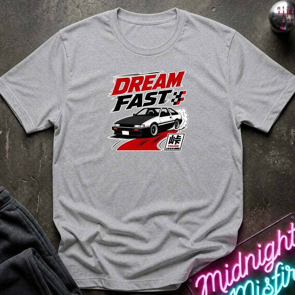 A heather gray short-sleeve graphic T-shirt laid flat on a dark textured surface. The shirt features a red, black, and white 'DREAM FAST' graphic with a car illustration.
