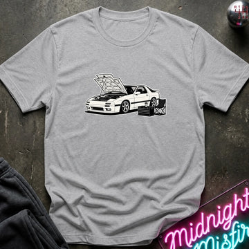 A light gray heather crewneck t-shirt featuring a black and white graphic of a sports car with its hood open, laid flat on a dark textured surface with a neon sign and dark pants partially visible.