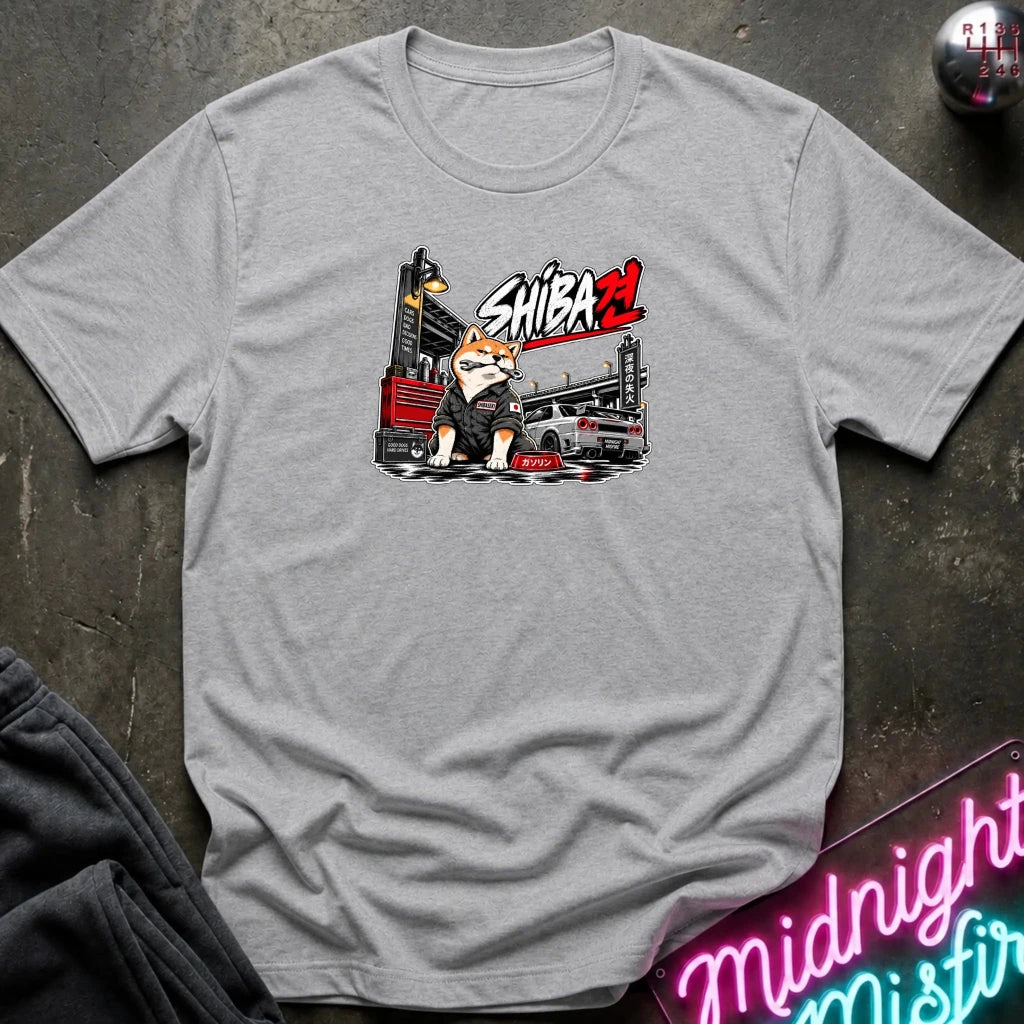 A light heather gray t-shirt laid flat on a dark textured surface, featuring a cartoon graphic of a Shiba Inu dog in a black suit with the text 'SHIBA' in red and white. 