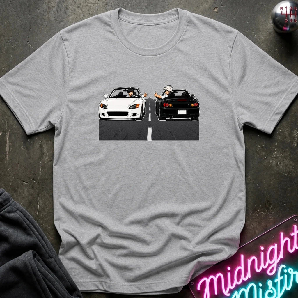 A heather gray short-sleeve t-shirt laid flat on a dark textured surface, featuring a graphic print of a white convertible and a black sports car facing each other on a road with a hand waving from the rear car.