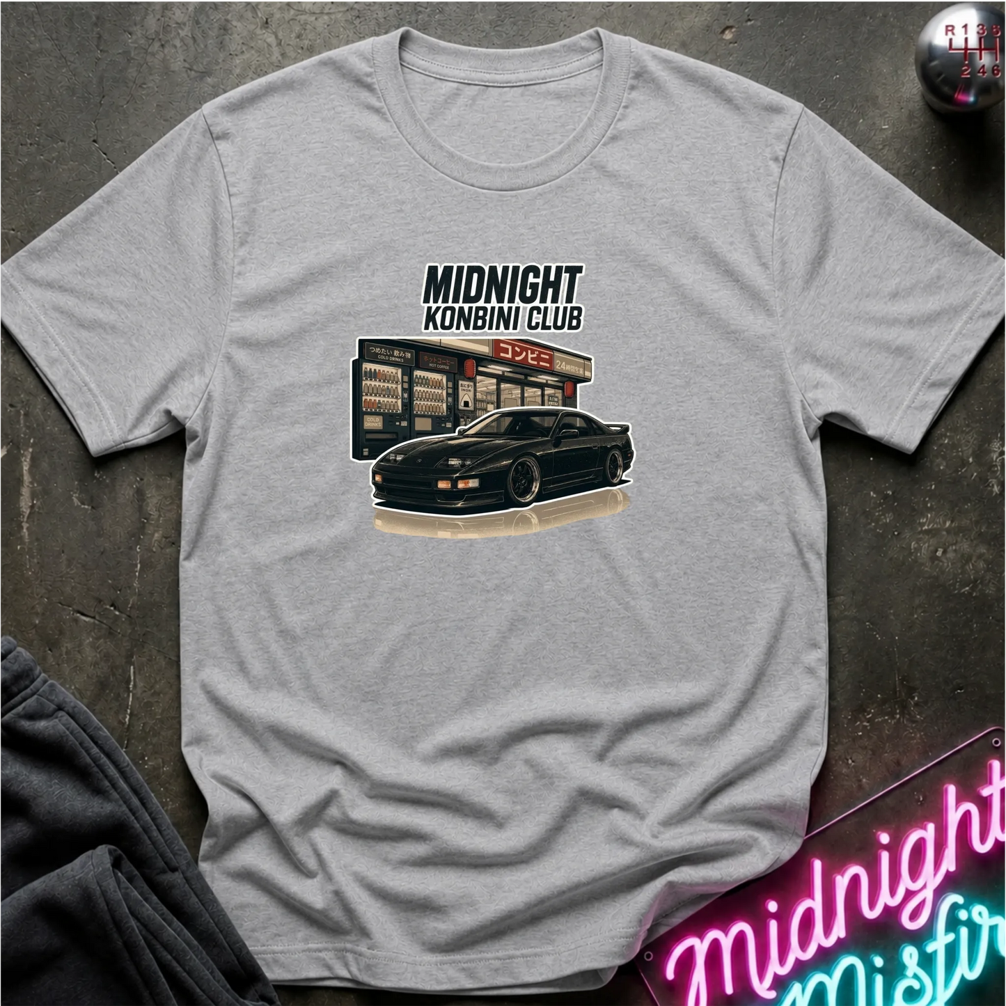 A light gray short-sleeve graphic t-shirt laid flat on a dark textured surface. The graphic features the text 'MIDNIGHT KONBINI CLUB' above an illustration of a black sports car parked in front of a convenience store.