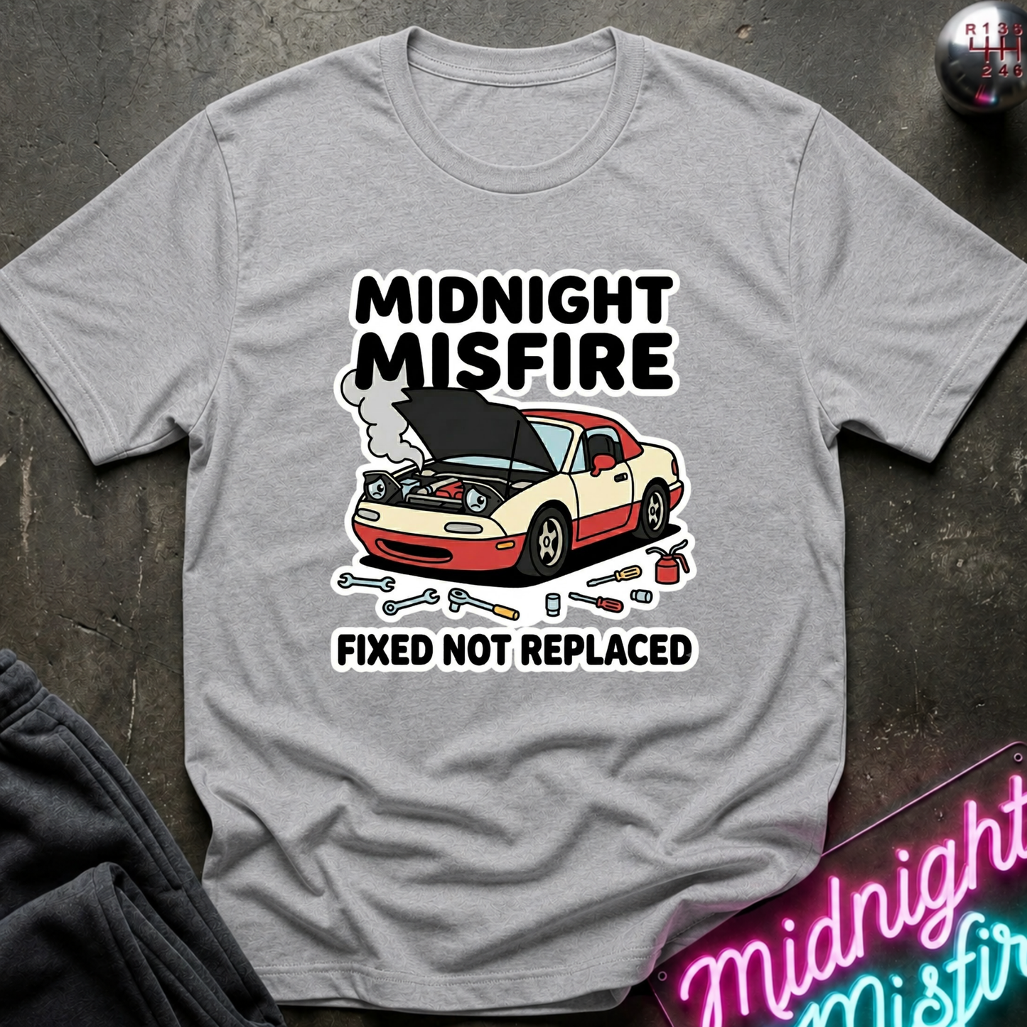 A light gray crewneck t-shirt featuring a graphic of a beige and red sports car with its hood open, surrounded by wrenches and the text 'MIDNIGHT MISFIRE' and 'FIXED NOT REPLACED'.
