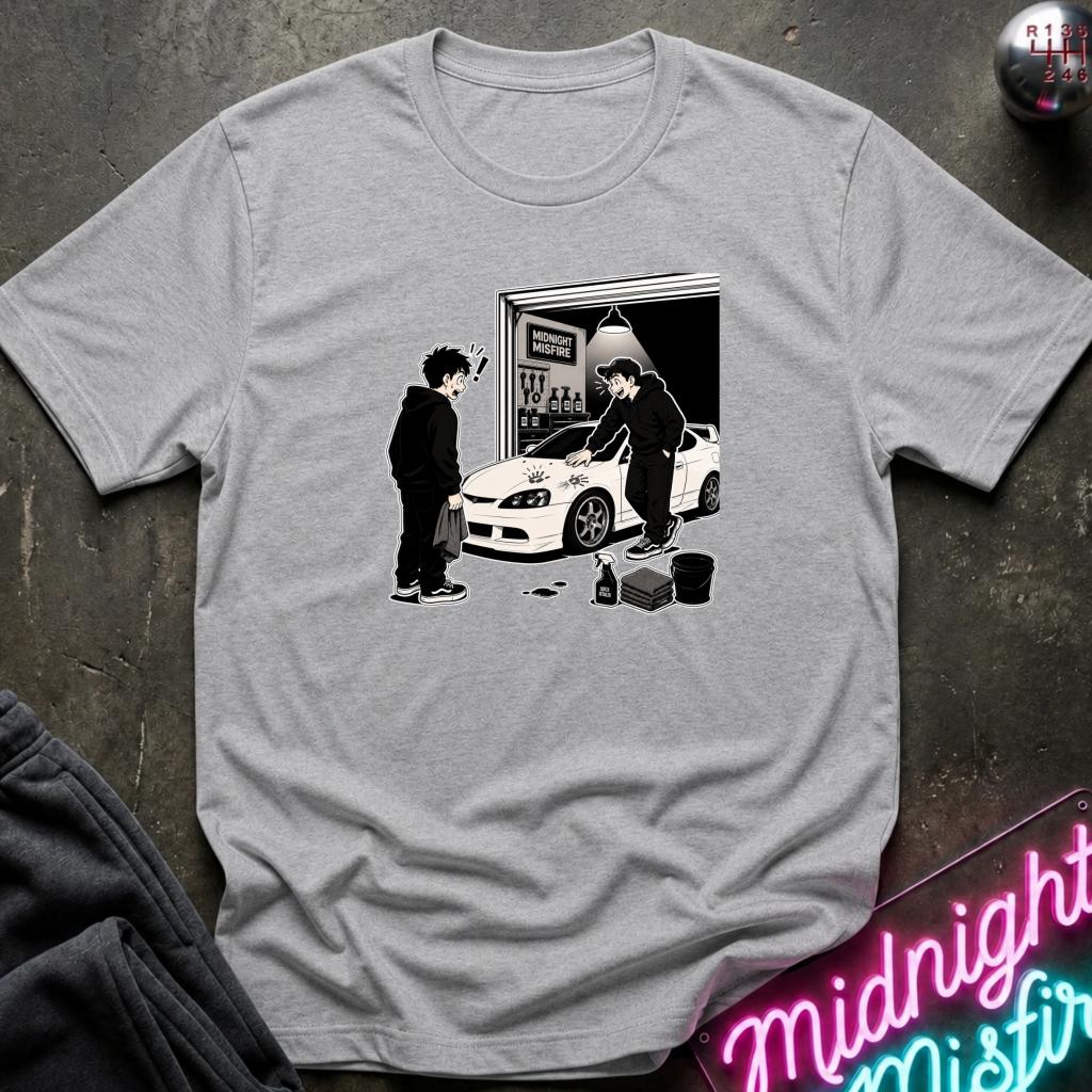 A light gray crewneck t-shirt with a black and white graphic print of a person washing a white sports car in front of a garage. The t-shirt is displayed flat on a dark textured surface with a neon sign and dark sweatpants partially visible.