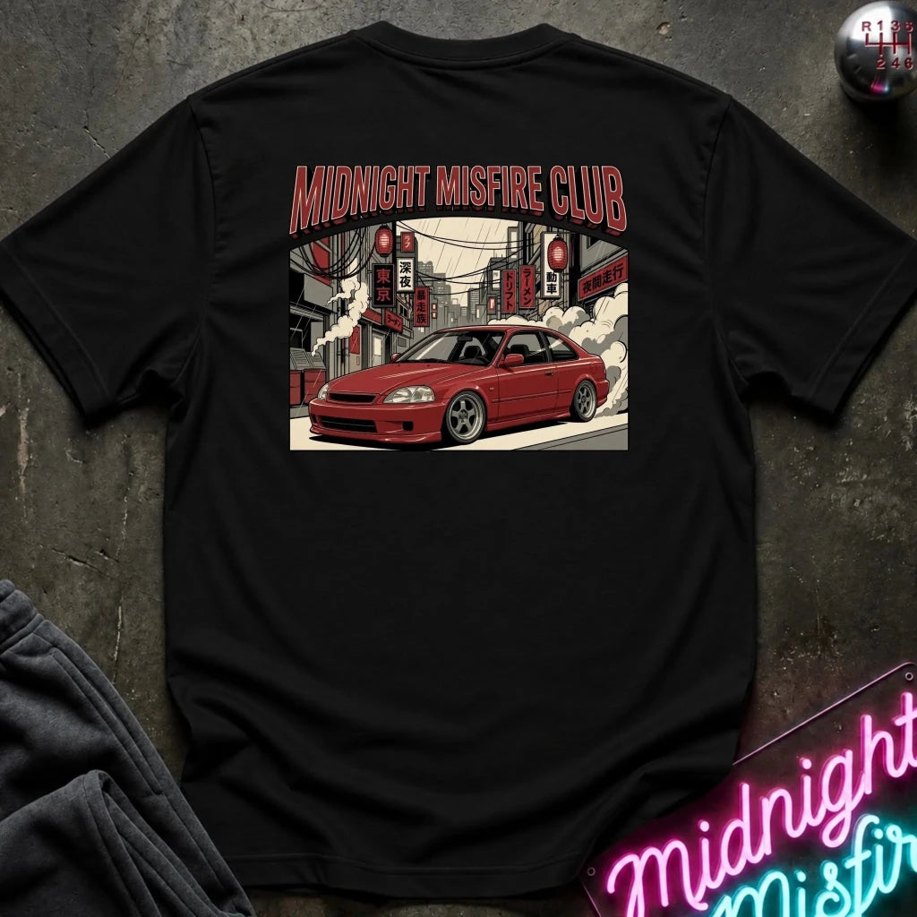 A black short-sleeve t-shirt laid flat on a dark textured surface, featuring a large graphic print on the back with the text 'MIDNIGHT MISFIRE CLUB' above an illustration of a red car in a city street scene. A neon sign and folded dark fabric are partially visible around it.