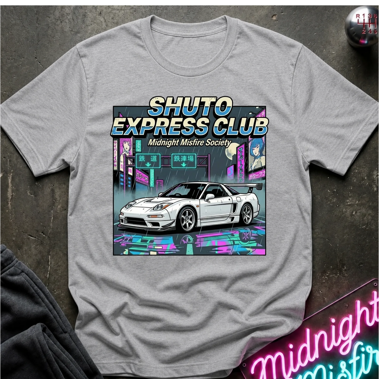 A heather gray short-sleeve t-shirt laid flat on a dark textured surface, featuring a large graphic print of a white sports car in a neon cyberpunk city scene with the text 'SHUTO EXPRESS CLUB' and 'Midnight Misfire Society'.