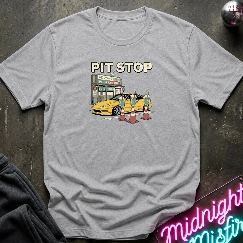 A heather gray crewneck t-shirt featuring a retro-style graphic of a yellow sports car at a Japanese convenience store with the text 'PIT STOP' above it, laid flat on a dark textured surface.