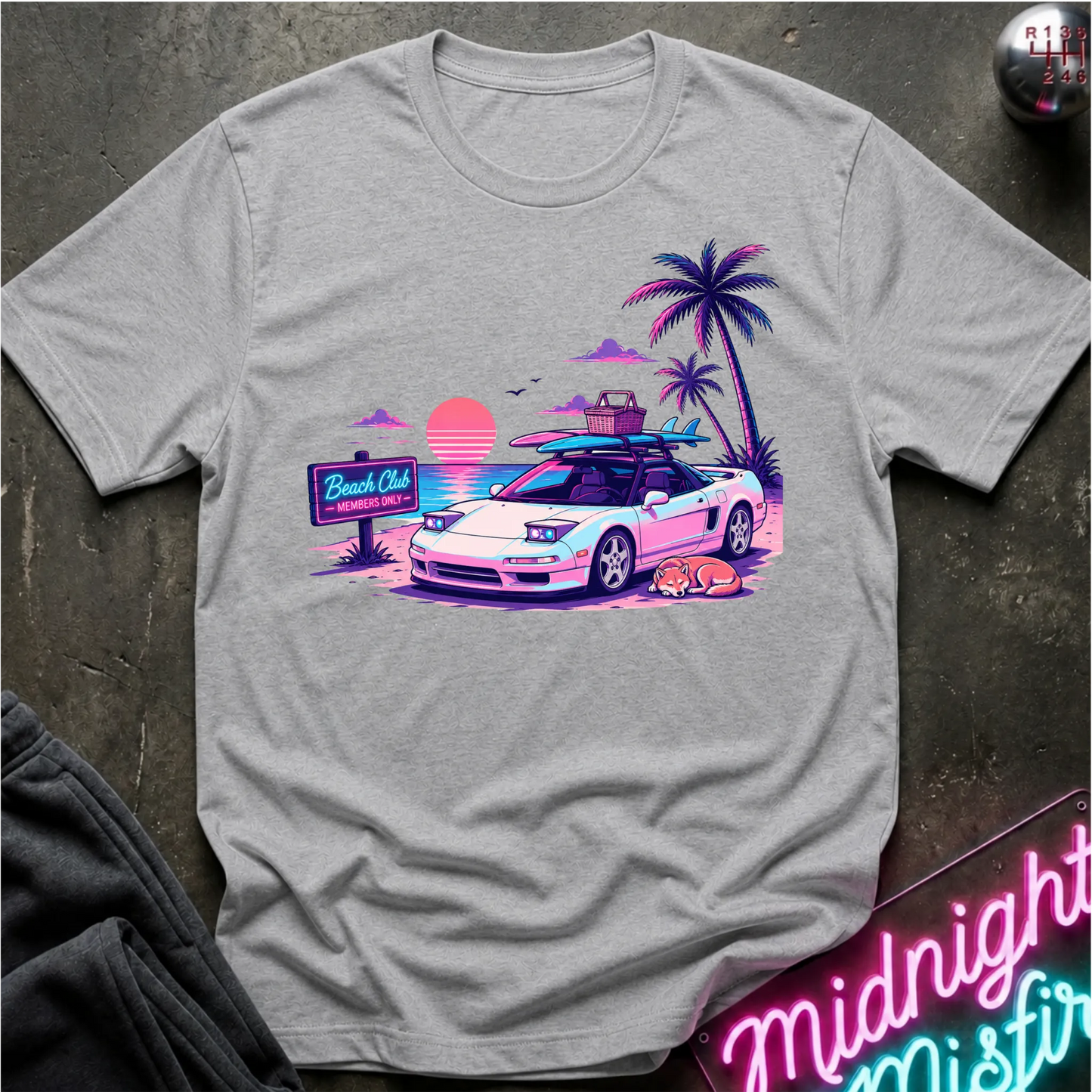 A light gray crewneck t-shirt featuring a colorful retro graphic of a white sports car on a beach with palm trees and a sunset. The shirt is laid flat on a dark textured surface with a neon sign and dark pants partially visible.