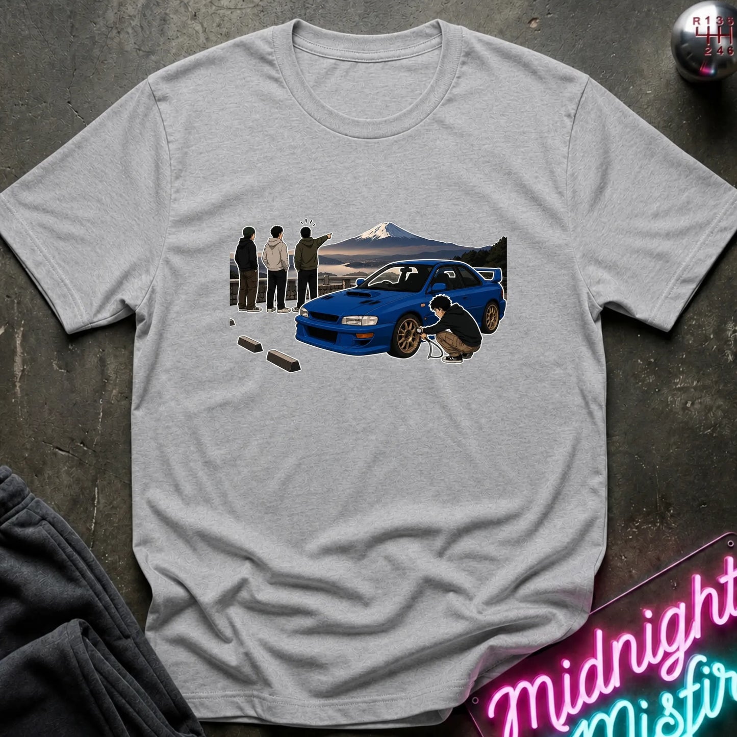 A light gray heather t-shirt laid flat on a dark textured surface, featuring a graphic print of a blue sports car in front of Mount Fuji with people and a dog. A neon sign reading 'Midnight Misfire' is partially visible in the bottom right corner.
