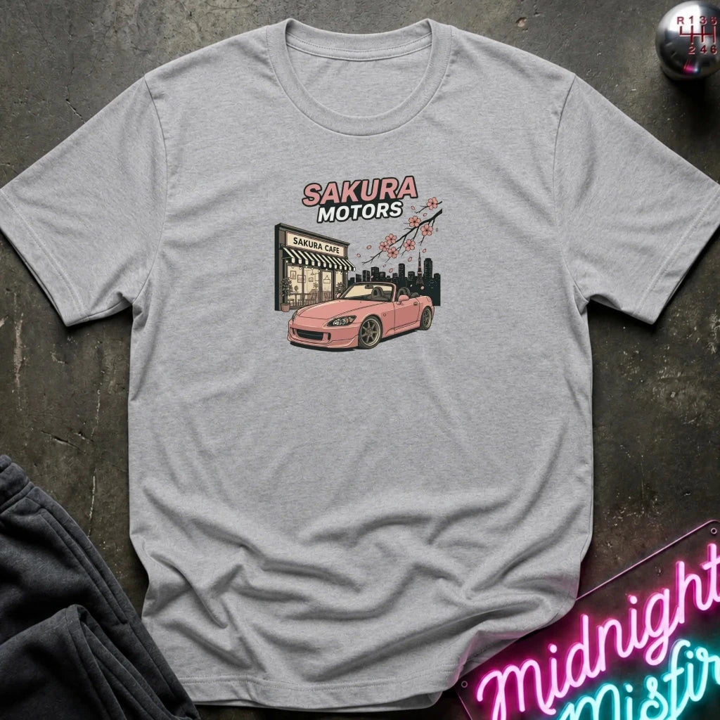 A light heather gray t-shirt laid flat on a dark textured surface, featuring a graphic print of a pink convertible car in front of a 'Sakura Motors' shop with cherry blossoms. A neon sign and dark clothing are partially visible in the background.