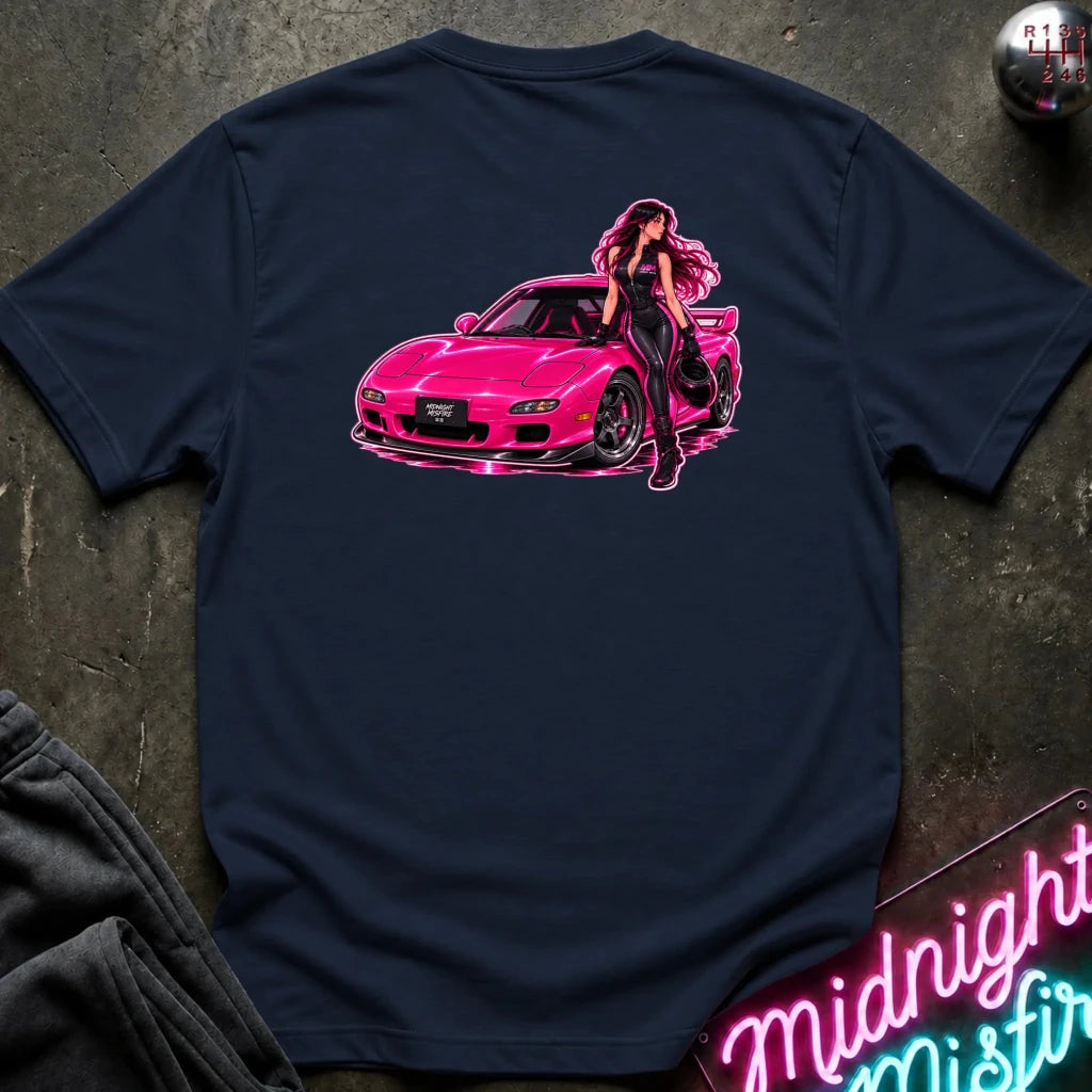 A navy blue t-shirt laid flat on a dark textured surface, featuring a graphic print of a pink sports car with a woman leaning on it. The shirt is styled with a dark sweatshirt, a gear shift knob, and a neon sign in the background.