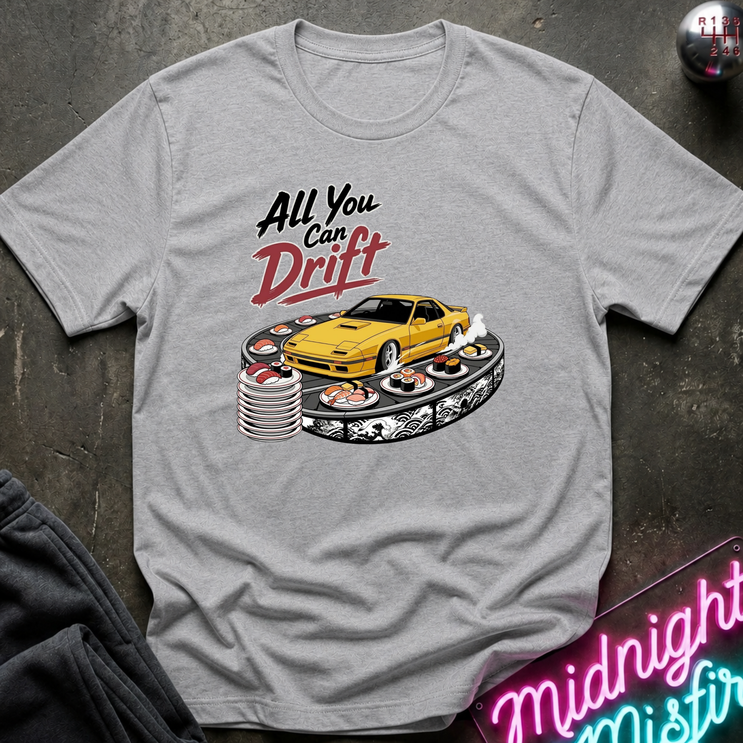 A light gray heather crewneck t-shirt featuring a graphic print of a yellow sports car drifting on a track with the text 'All You Can Drift' in black and red. The shirt is displayed flat on a dark textured surface with a neon sign and dark pants partially visible.