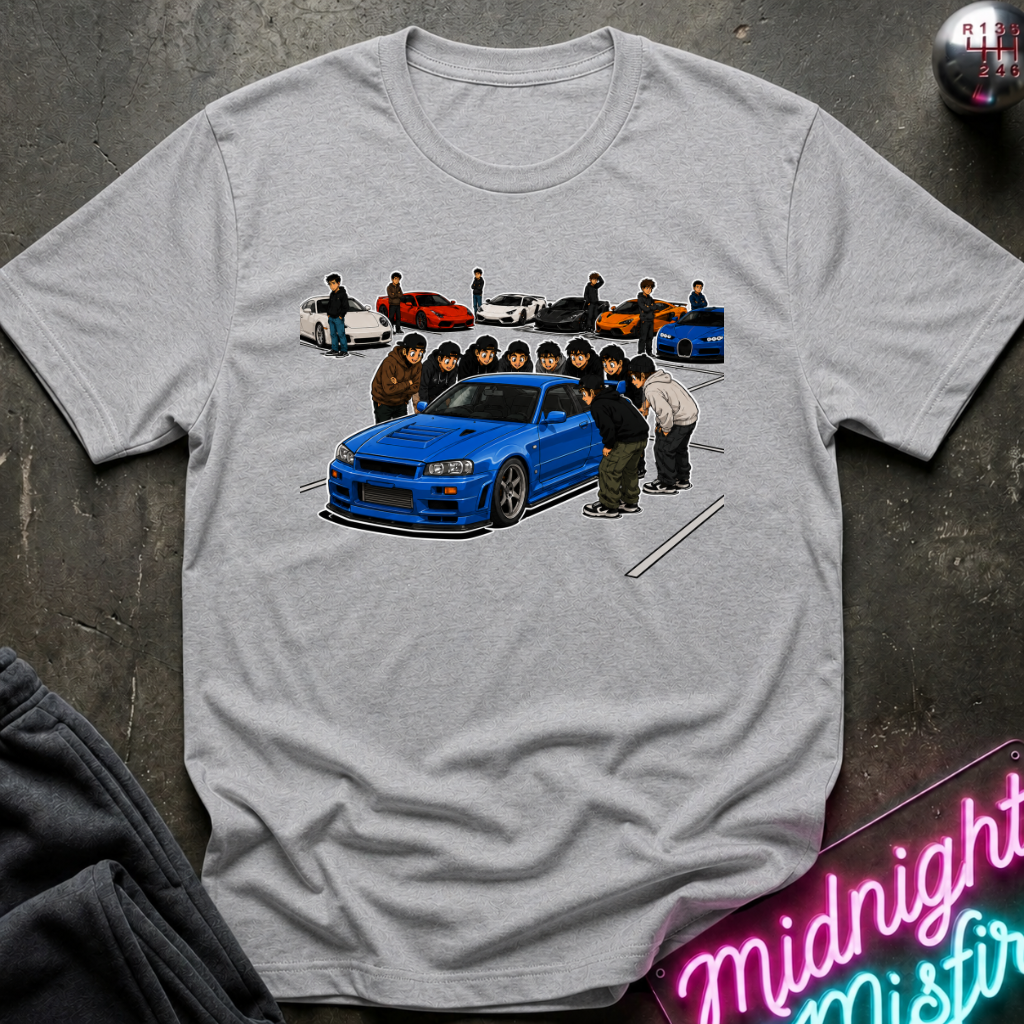 A light gray heather t-shirt laid flat on a dark textured surface, featuring a graphic print of a blue sports car with a group of anime-style characters standing around it. A neon sign reading 'Midnight Mistir' is visible in the bottom right corner.