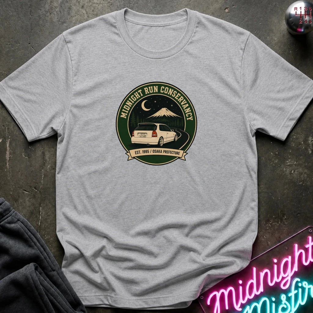 A light heather gray short-sleeve t-shirt laid flat on a dark textured surface. The center of the shirt features a circular graphic with a mountain, moon, and a white SUV, with the text 'Midnight Run Conservancy' and 'EST.